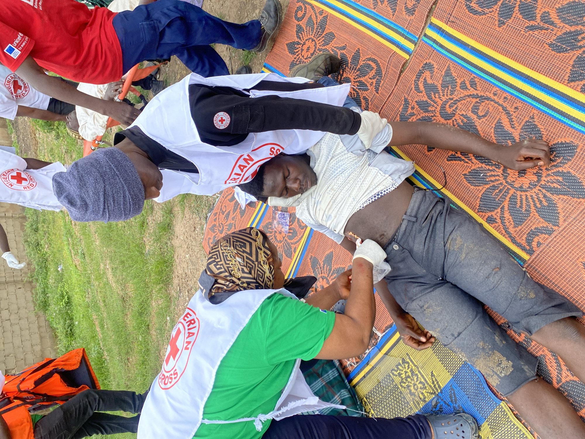 In Kawo, Nigeria, a man lies on a mat on the ground, seemingly receiving medical attention. He is dressed in a light-colored, striped shirt and dark grey, knee-length pants that are soiled with dirt. His abdomen is exposed, and a person wearing white gloves is attending to him. This caregiver is wearing a green shirt and a white vest with the Nigerian Red Cross emblem. Another individual, also in a white vest with the Nigerian Red Cross emblem, is kneeling beside the injured man, wearing a grey beanie and black long-sleeved shirt. A red and orange life vest is visible near the top left of the frame, and further back, two other people in red shirts with a UNICEF logo are also present. The ground appears to be a mixture of grass and dirt. The overall scene suggests a medical emergency or first aid situation.