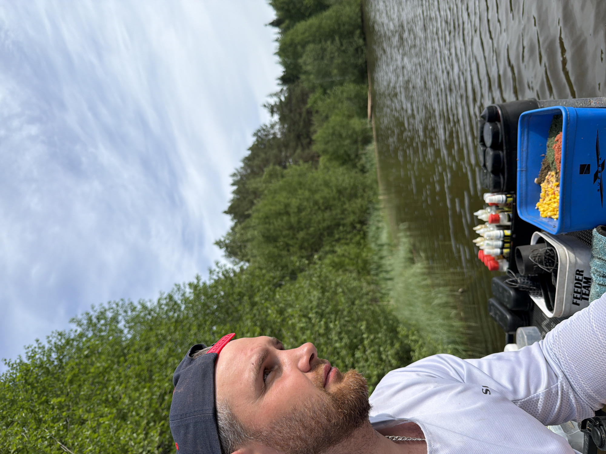 A man is seated on the edge of a body of water, possibly a river or lake. He is wearing a white shirt, a dark cap, and a silver chain. Next to him are fishing supplies, including a blue bucket containing bait and various containers with red and white caps. The backdrop consists of trees and a cloudy sky. There is no explicit indication of location within the city of Prikamskii, Russia.