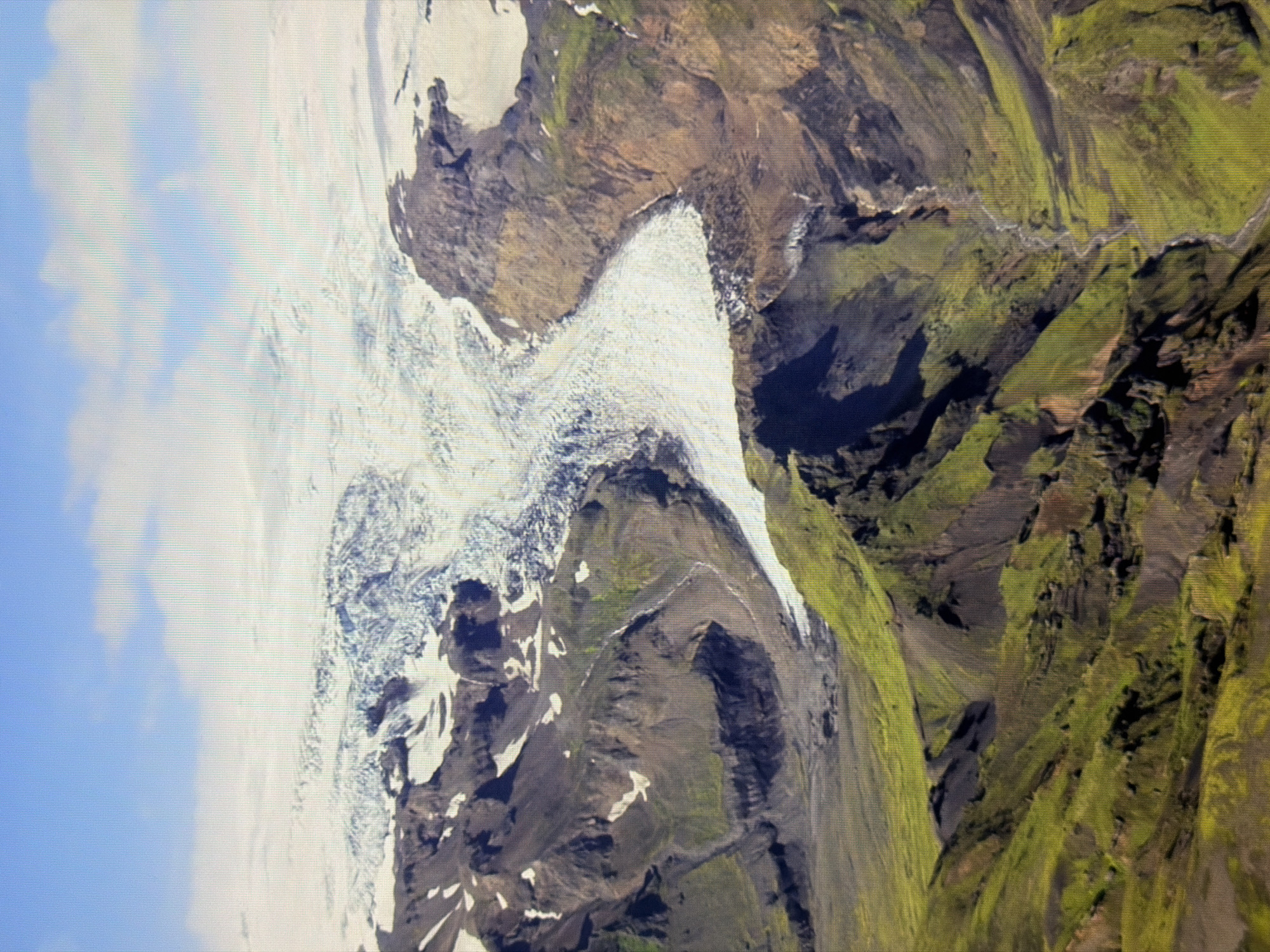 The image is an aerial view of a glacial landscape. A large, white glacier dominates the upper portion, snaking down between rugged, dark brown and green mountains. The mountains show signs of volcanic activity with dark ash deposits mixed with green vegetation. A river or meltwater channel carves its way through the landscape, visible in the lower right portion of the image. The sky above is blue with scattered white clouds. There are no people, structures, or signs of urban life visible. The scene depicts a remote and natural environment. The location is not in Kyiv, Ukraine.