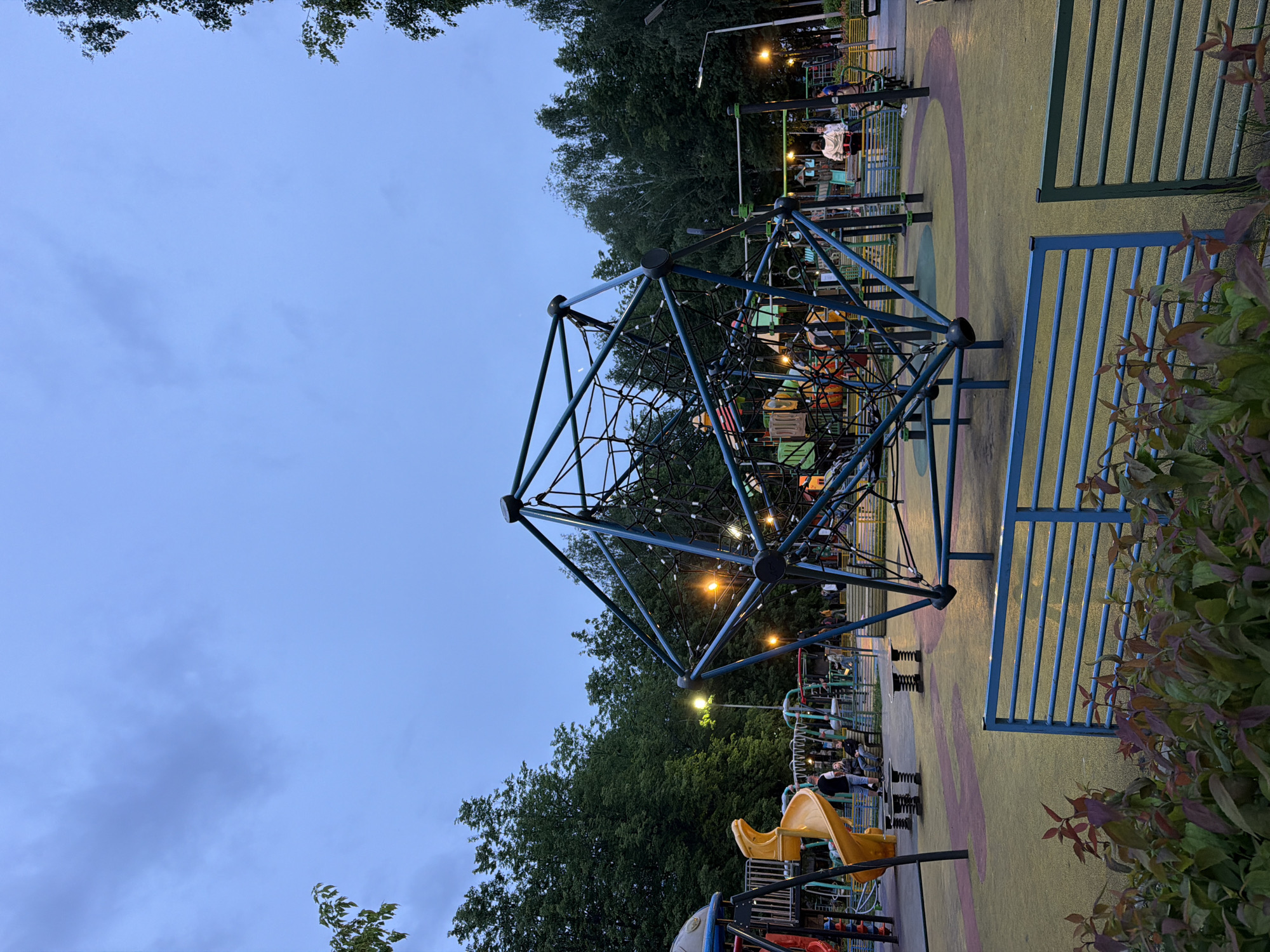 The image shows a playground in Dolgoprudny, Russia. The main structure is a large, geometric climbing frame made of interconnected blue metal rods and ropes. In the background, there are other playground elements including slides and spring riders. Several people are visible, some sitting on benches and others on the playground equipment. Street lights illuminate the scene, indicating it is dusk or evening. The playground is surrounded by trees and shrubbery. The ground is covered with a rubberized safety surface, colored with various patterns. A blue metal fence is on the right side of the image. The sky is overcast with a light blue hue.