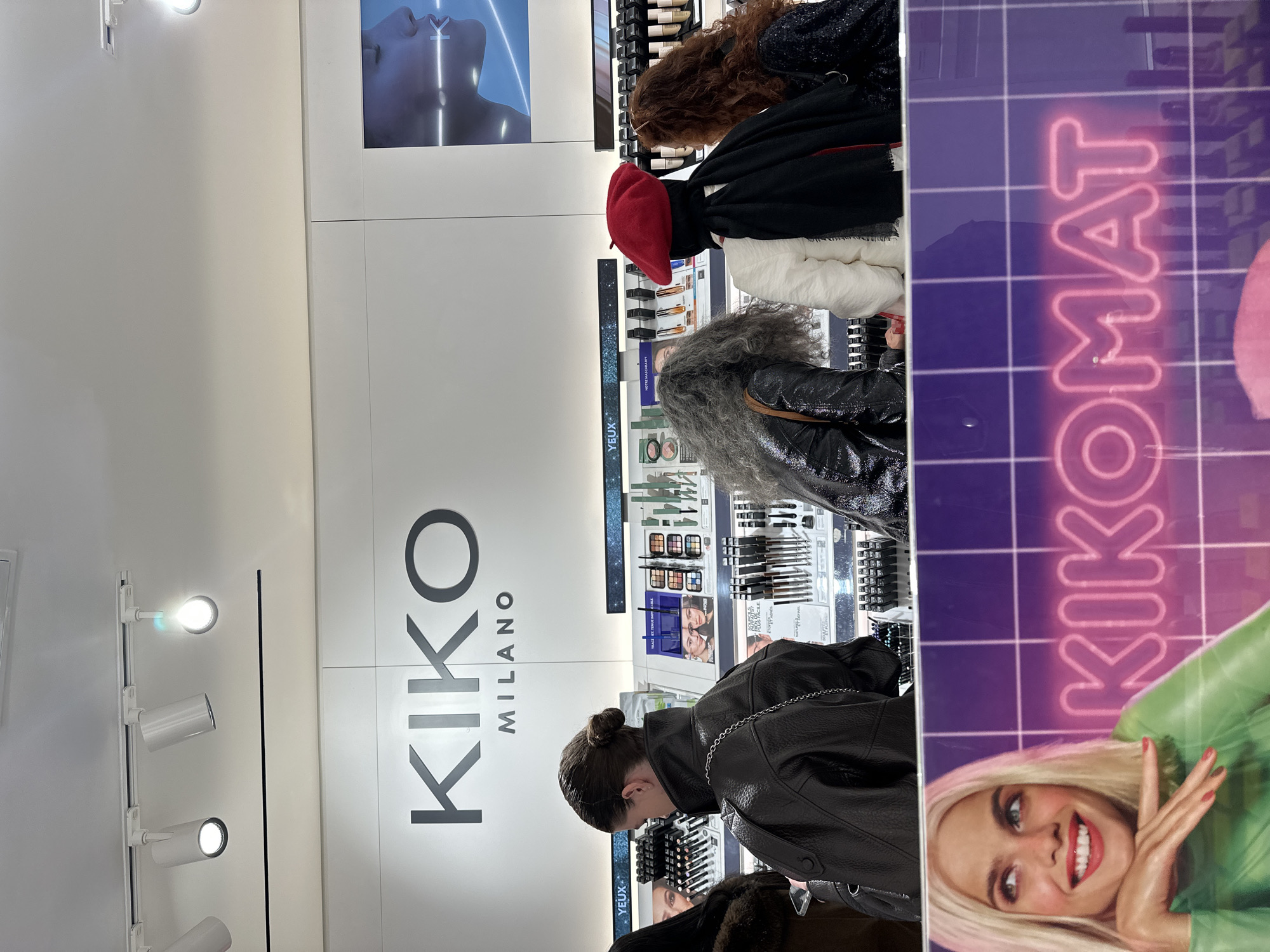 The image shows the interior of a KIKO Milano store in Paris, France. Customers are browsing the makeup displays. The store's branding is visible with the logo "KIKO MILANO" on the wall. Various makeup products are on display. A promotional poster with "KIKOMAT" text and an image of a woman is seen on the side. There are overhead lights and a mannequin image. The people are focused on the makeup products and the environment is that of a retail cosmetics store.