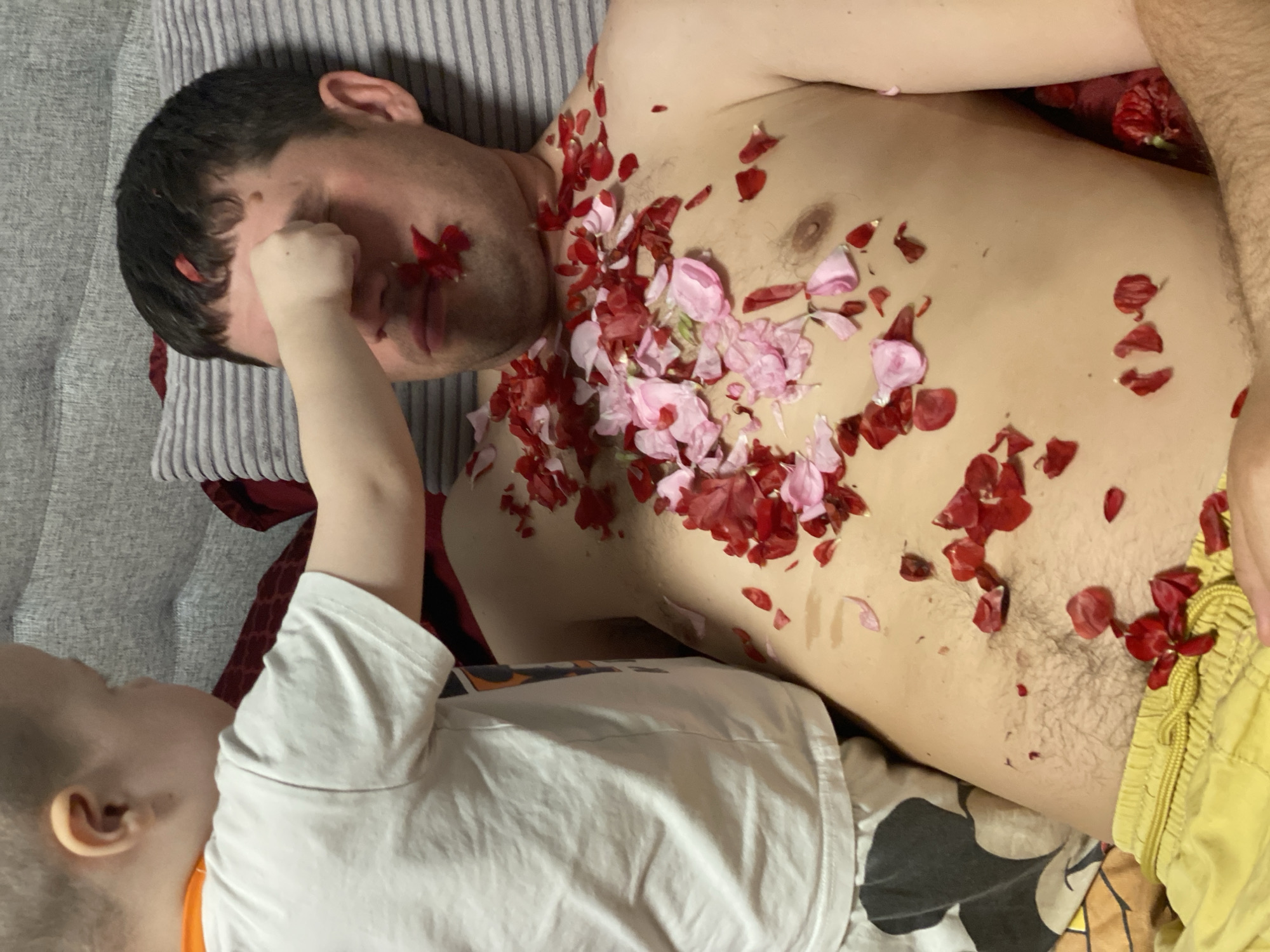 This indoor image, likely set in a home in Sochi, Russia, captures a playful and tender moment between an adult male and a young child. The adult male, partially shirtless, is lying down, possibly resting or sleeping, on a striped gray and white pillow. His torso is adorned with numerous red and pink flower petals, which are also scattered on his face, including on his closed eyes and mouth. He is wearing yellow shorts.
A young child, seen from the chest up, is positioned next to the adult, facing him. The child is wearing a white t-shirt with a visible orange collar. Their left hand is extended towards the adult's face, seemingly placing more petals or gently interacting. The child's bed or blanket, featuring a cartoon-like pattern in black, white, and brown, is visible beneath them.
The scene conveys a sense of lightheartedness and affection, with the child engaged in a playful activity involving the flower petals and the adult appearing to be a willing, albeit possibly sleepy, participant. The emotions evoked are primarily tenderness, playfulness, and relaxation. No specific time of day is discernible, and there is no visible text in the image.