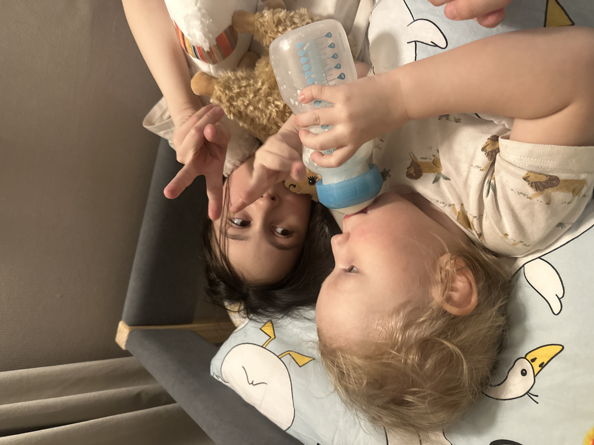 The image depicts two individuals, likely a child and an adult or older sibling, lying on a bed. The child, with fair hair, is positioned on a light blue blanket with a goose motif and appears to be drinking from a baby bottle. The other individual, with dark hair, is making a "peace" sign with their fingers. They are holding a stuffed llama toy. The location cannot be determined to be in Votkinsk, Russia from the image alone. The environment appears to be indoors, likely a bedroom. A portion of a gray headboard is visible.