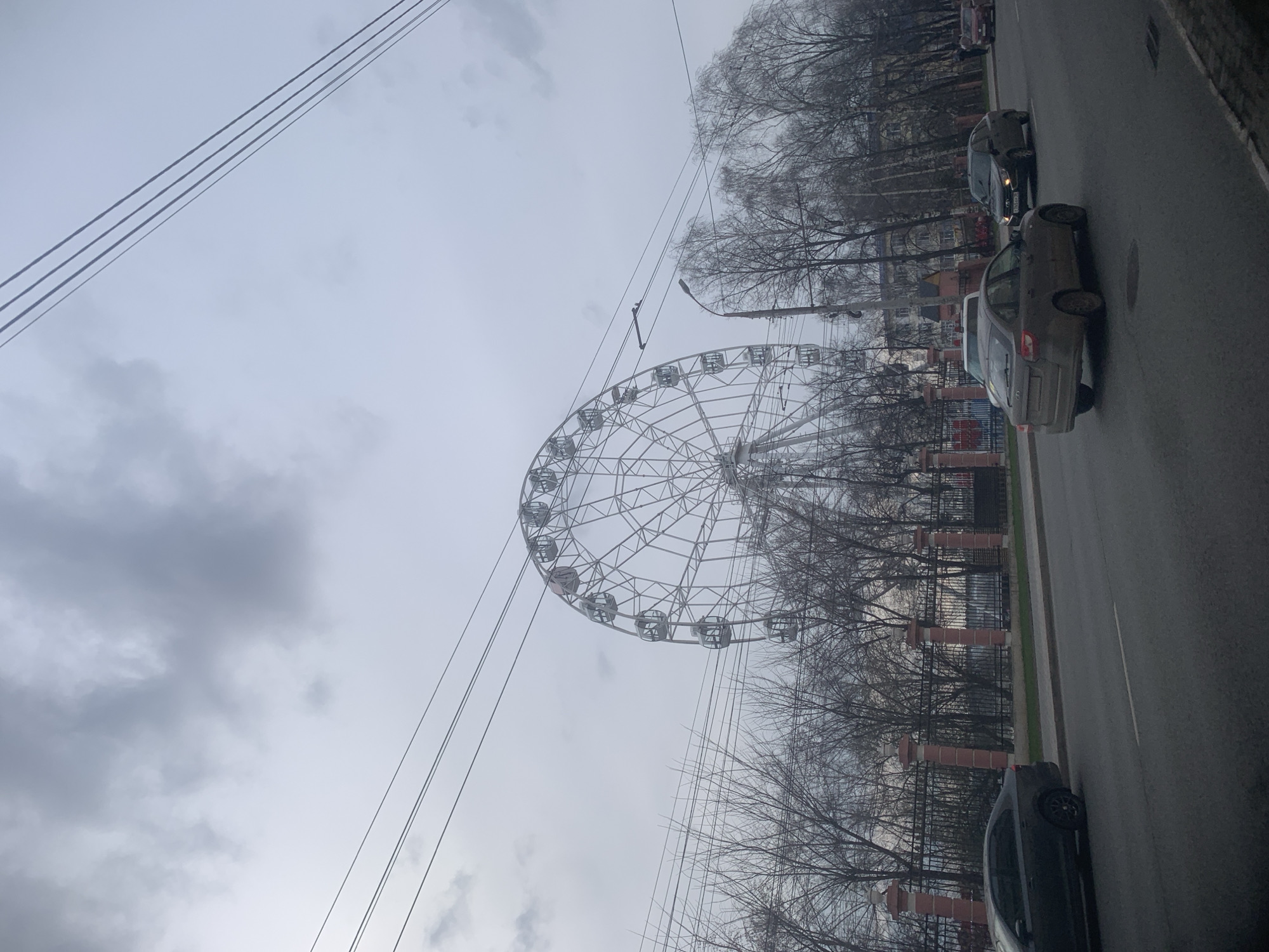 The image depicts a street scene in Izhevsk, Russia, featuring a Ferris wheel prominently positioned against a cloudy sky. The Ferris wheel appears to be located within a park or recreation area, partially obscured by bare trees. A road runs along the right side of the frame, with parked cars lining the curb. Overhead power lines extend across the upper left portion of the image. The overall impression is of a gray, overcast day in an urban environment.
