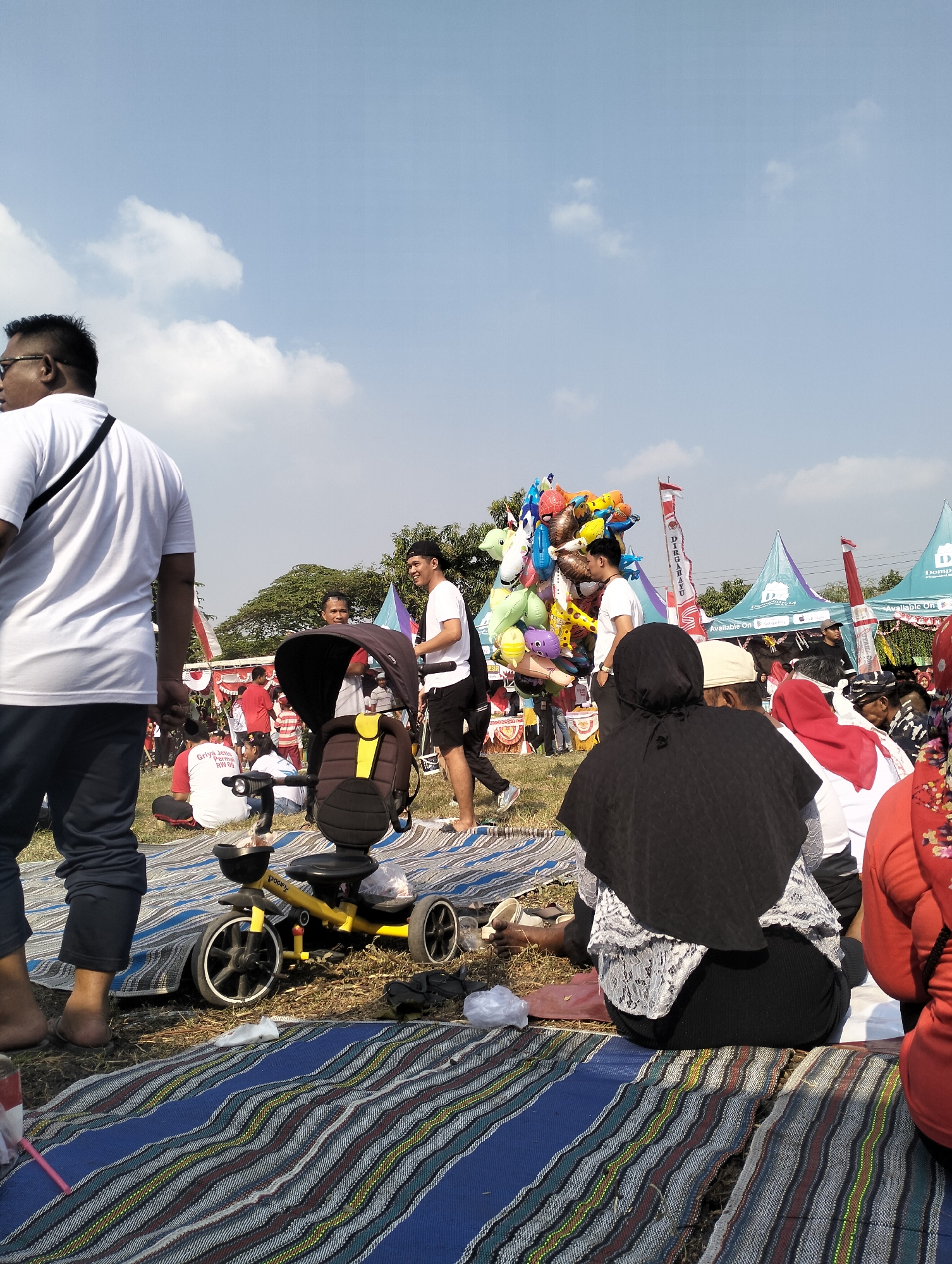 The image depicts an outdoor gathering, likely a festival or community event, taking place on a sunny day in Jetis, Indonesia. In the foreground, a person wearing a white t-shirt and dark pants is standing, viewed from the back and left side. To their right, a yellow children's tricycle with a dark brown canopy is parked on a striped mat. Several people are seated on mats in the midground, facing away from the camera. One individual is wearing a black headscarf and a patterned top. Another person wears a red head covering. A cluster of colorful balloons, including characters like Spiderman and a giraffe, are held aloft by a person standing in the midground, partially obscured by the balloons. In the background, tents are set up, some bearing logos and text, suggesting commercial or organizational presence. Flags and banners are visible. Trees and foliage form the backdrop. The ground appears to be covered in grass and dirt, with people sitting on various mats. The sky is clear with scattered white clouds.
