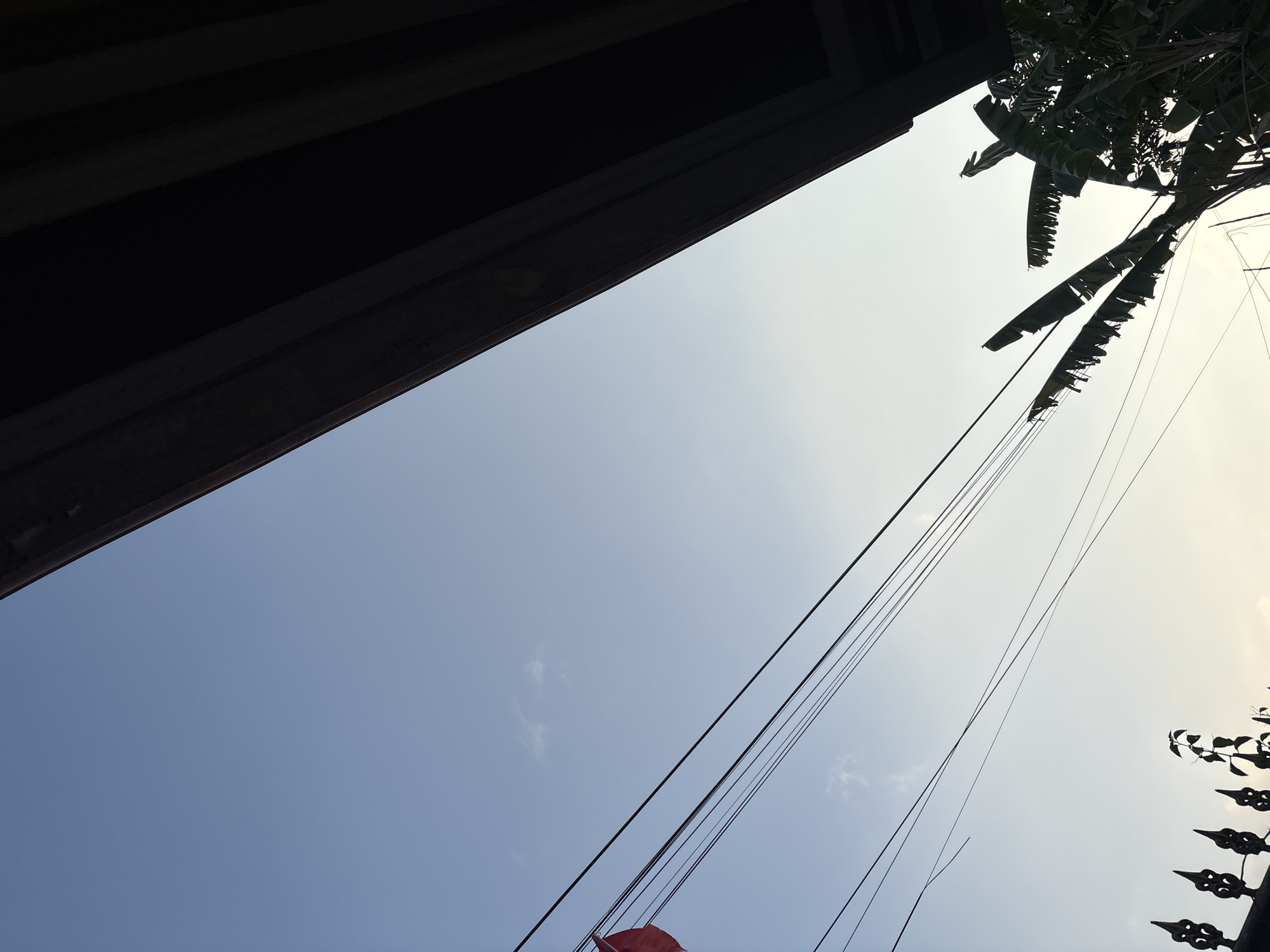 The image is an outdoor, low-angle shot looking up at the sky from within a city. A dark, textured roof overhang dominates the top right portion of the frame, casting a shadow. Below this, a clear blue sky stretches across the majority of the image, with a few faint clouds visible. Several black electrical wires are stretched diagonally across the sky, originating from the left and extending towards the right. In the lower left corner, a black metal fence with ornate spikes is partially visible, with a small plant growing above it. To the right of the fence, a silhouette of banana tree leaves is visible against the lighter sky. A small fragment of a red object, possibly a flag or banner, is visible on the far left, attached to one of the wires. The overall scene suggests an urban or suburban environment in Palimanan, Indonesia, likely during the late afternoon or early morning, given the quality of light. There are no people or visible actions depicted in the image.