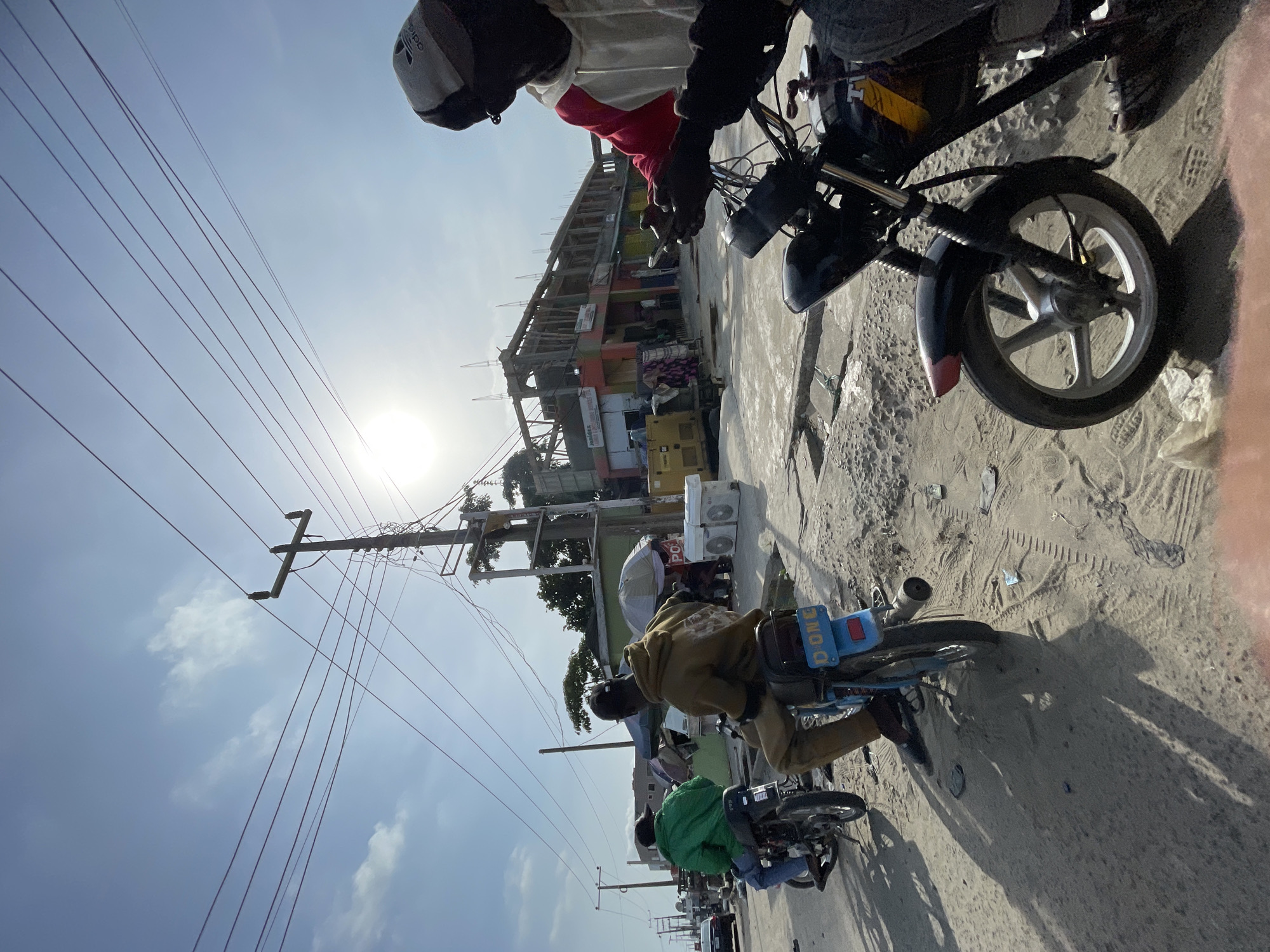 Several motorcycles are on a sandy road in Ikorodu, Nigeria. In the foreground, a person in a white and red jacket rides a motorcycle. Further down the road, two people on motorcycles wear green and brown jackets. Power lines crisscross the sky above the scene. Buildings line the street. The sun is bright.