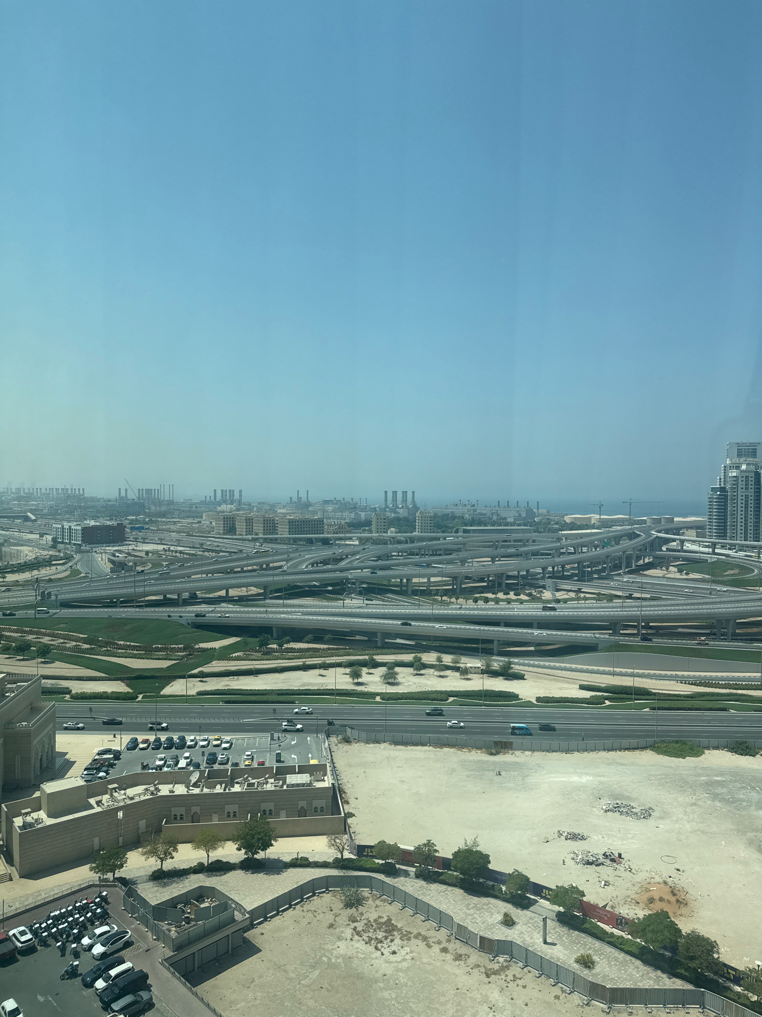 This is an aerial view of a complex highway interchange in Dubai, United Arab Emirates, under a clear blue sky. The interchange features multiple levels of elevated roads and ramps, with traffic moving on them. In the foreground, a building with an architectural style featuring arched windows and a flat roof is visible, with a parking lot filled with cars and motorcycles adjacent to it. Further in the background, more buildings and industrial structures can be seen, along with the sea in the distance. The landscape around the interchange is a mix of green spaces and arid, undeveloped land with scattered piles of debris.