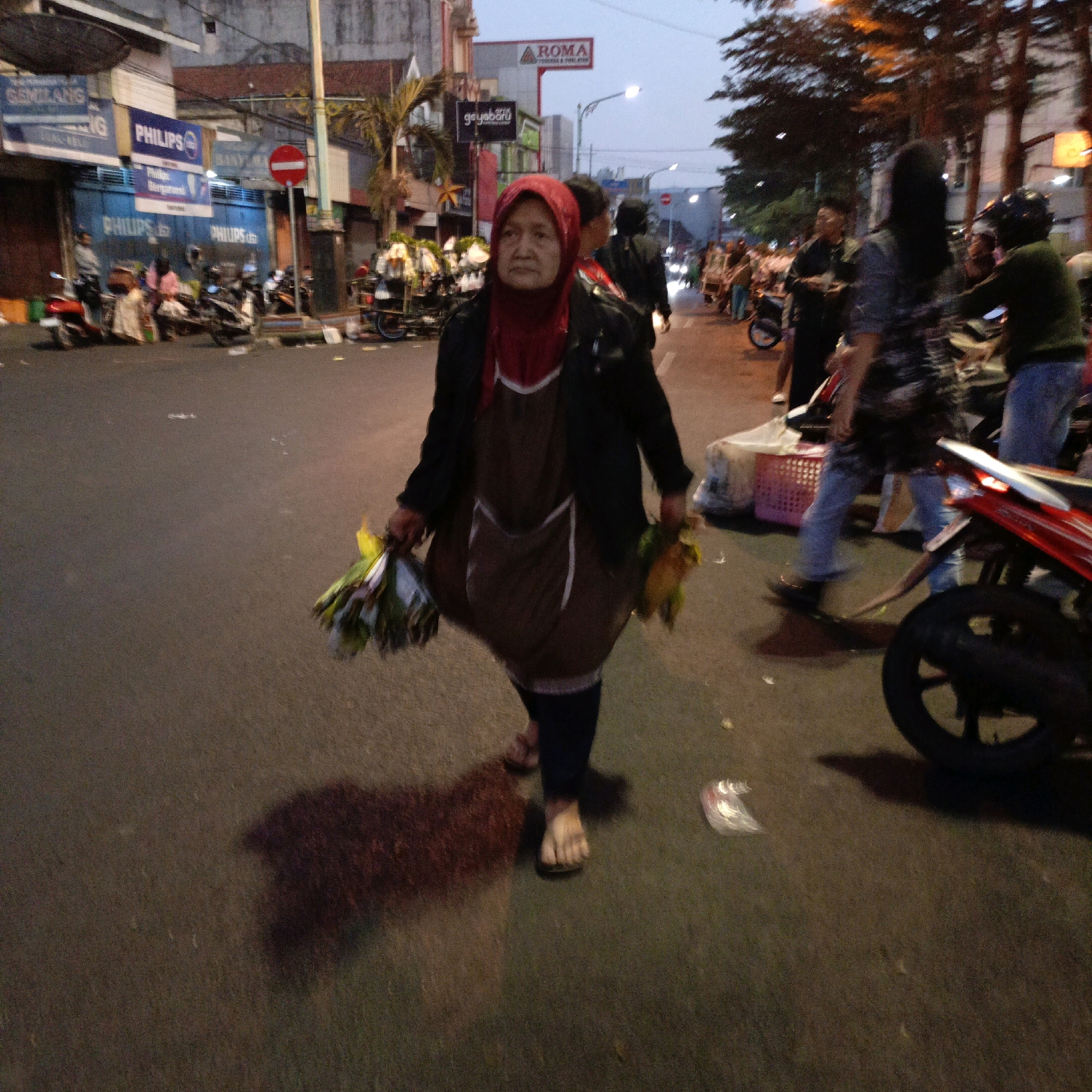 The image depicts a street scene in Salatiga, Indonesia, likely during the late afternoon or early evening due to the lighting conditions. In the foreground, a woman wearing a red hijab and a brown apron walks towards the camera, carrying two bunches of what appear to be vegetables or flowers, one in each hand. She is dressed in a black jacket over a brown garment. Her shadow is cast on the asphalt road behind her.
The background shows a street lined with buildings and trees. Signs for businesses like "PHILIPS" and "ROMA" are visible on the buildings. Several motorcycles are parked or moving on the street, and other people are present, some also appearing to be on motorcycles. A red motorcycle is prominently featured in the mid-right of the image. The overall atmosphere suggests a bustling urban environment.