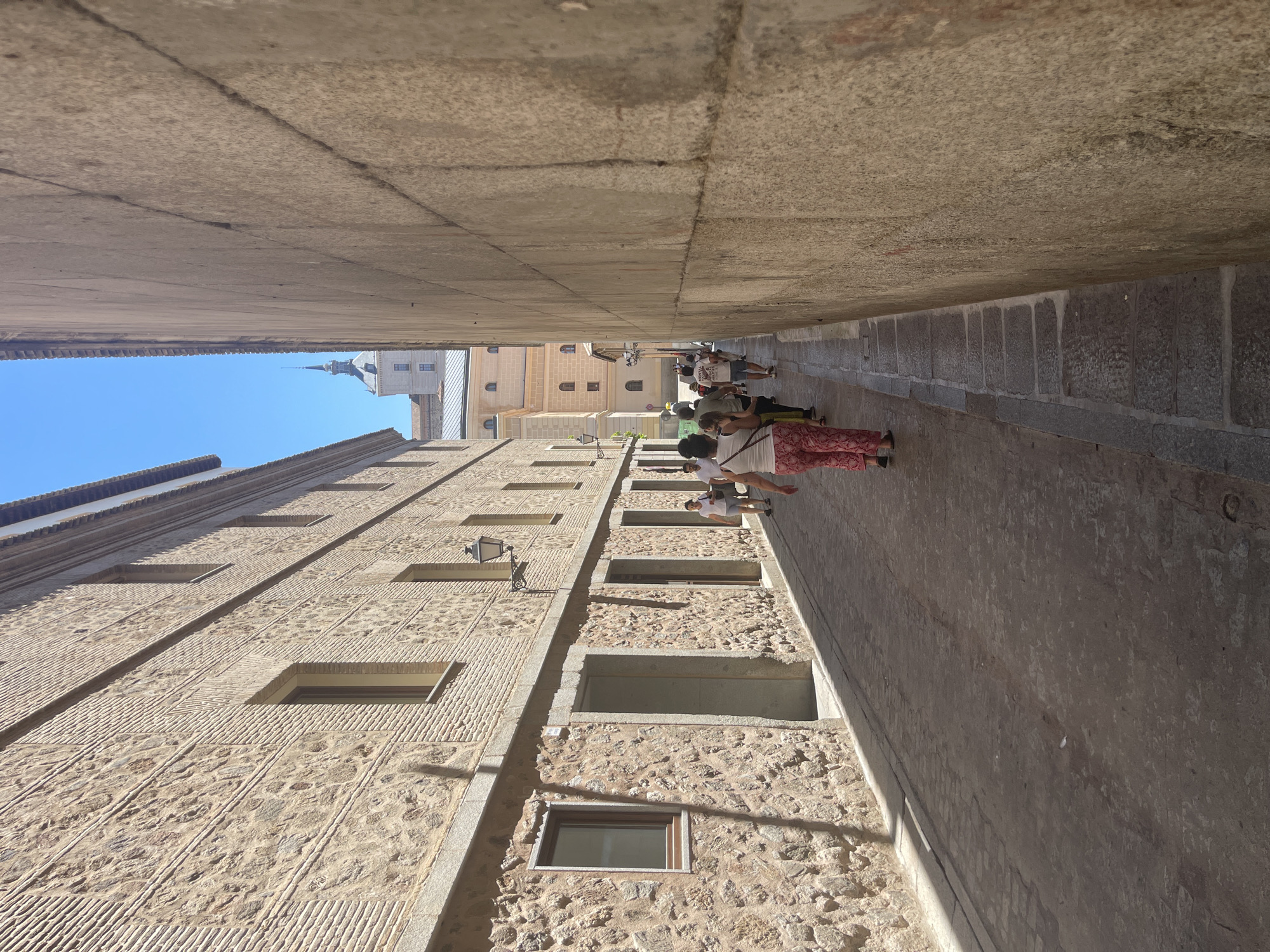 The image depicts a narrow, cobblestone street in Toledo, Spain, with tall, stone buildings flanking both sides. The street is cast in shadow, with sunlight illuminating the upper portions of the buildings and the sky above. Several people are walking down the street, appearing to be tourists. On the left, a building exhibits a facade of alternating stone and brickwork, with several windows and doorways visible. A decorative streetlamp is attached to the wall. On the right, a massive stone wall dominates the view, its rough texture and cracks evident. In the distance, at the end of the street, a prominent tower with a spire rises against the clear blue sky, suggesting a significant architectural landmark within the city, likely a church or cathedral. The overall impression is one of historical charm and a warm, sunny day in a European city.