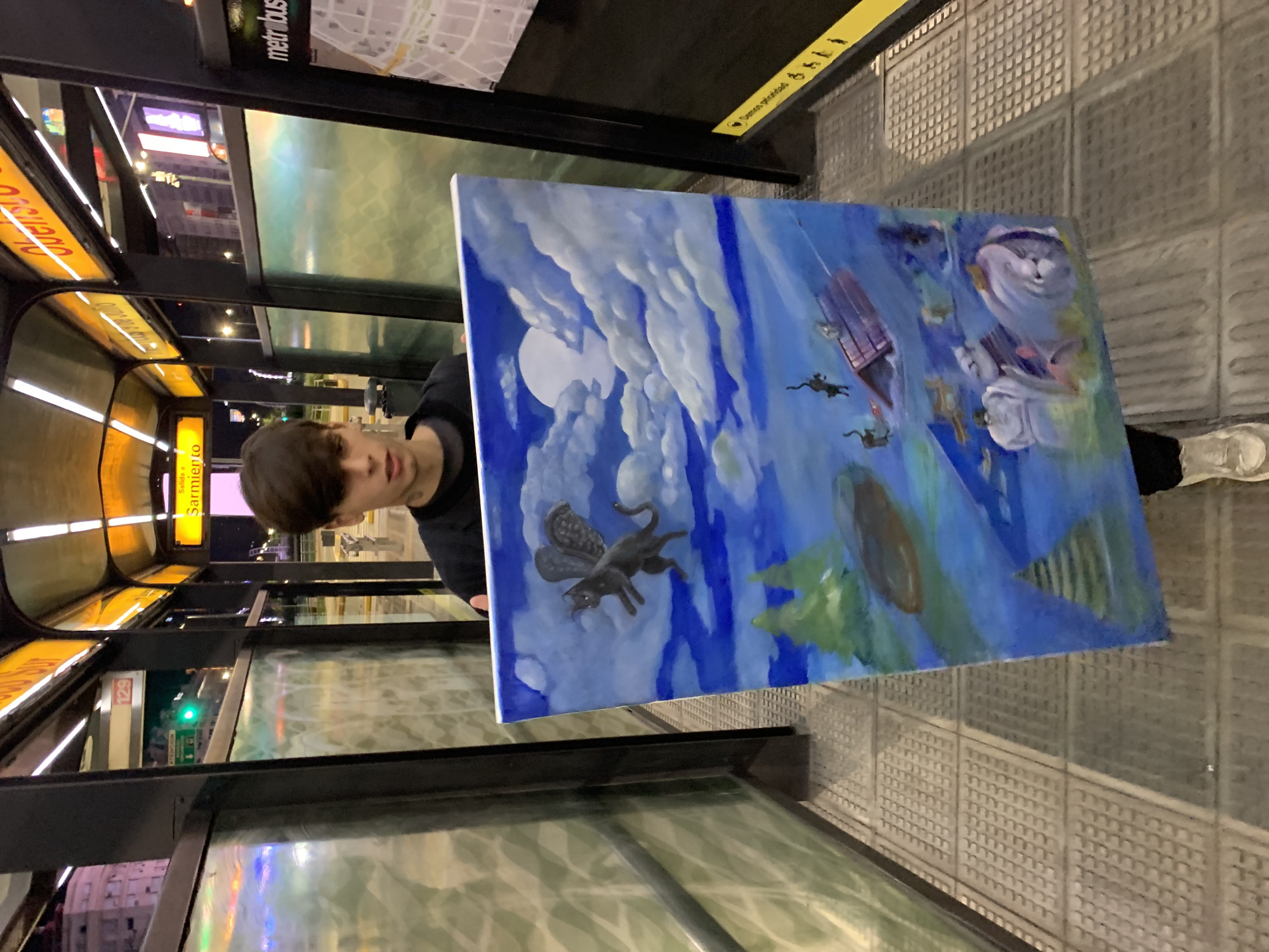 A person with dark hair holds a painting in front of a subway platform at a Buenos Aires Metro station, Argentina. The station name "Sarmiento" is visible. The painting depicts a surreal scene with clouds, a full moon, a winged black cat, a building, and a cartoon-like cat face. The subway platform is tiled with a patterned surface. A partially visible shoe of the person holding the painting is visible. The subway station has yellow illuminated signage. A "metrobus" map is visible above and behind the person holding the painting.