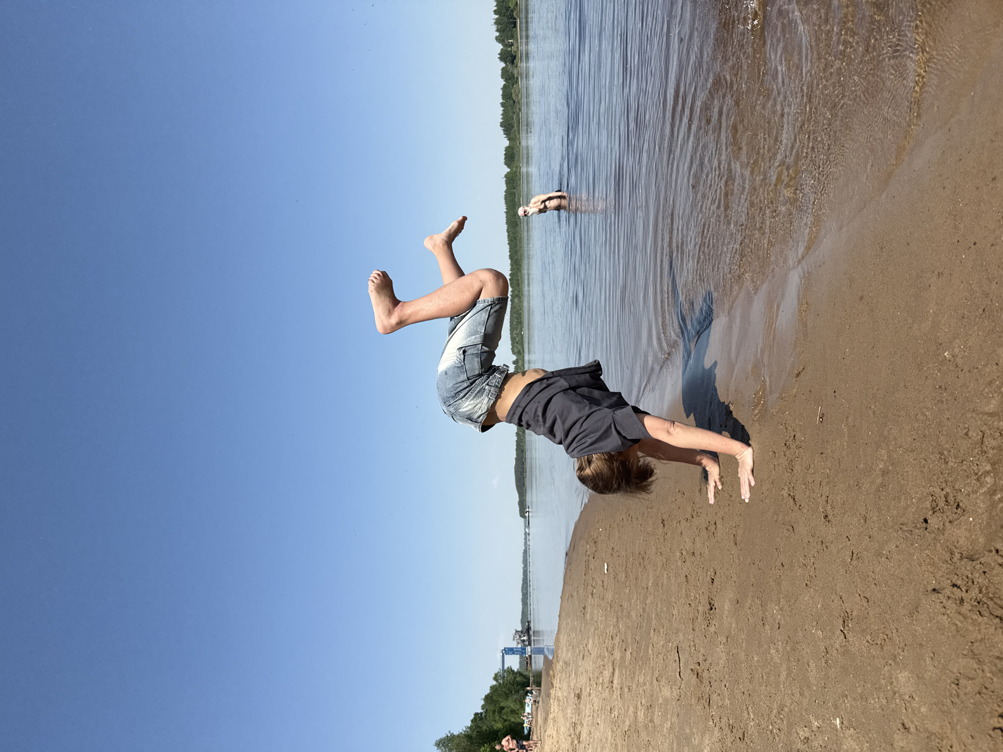 A medium shot depicts a person performing a handstand on a sandy beach, with their body inverted against a bright blue sky. The individual, wearing denim shorts and a dark grey shirt, is positioned with their feet pointed upwards towards the top left corner of the frame. Their hands are planted on the sand, supporting their weight.
In the background, a body of water stretches horizontally across the scene. There is a figure swimming in the water, located centrally in the background. The water extends to meet the treeline on the horizon.
In the bottom left of the frame, there is a crane structure, a line of trees and people standing on the sand. The location is the city of Unknown, Unknown.