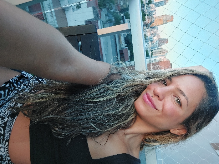 A woman is depicted in a close-up selfie taken on a high-rise balcony in Buenos Aires, Argentina. She is smiling directly at the camera, with a head and upper torso visible. She has long, wavy hair with prominent blonde streaks, dark eyes, and pink lipstick. A small metallic stud piercing is present above her upper lip. She wears a black crop top and black and white patterned shorts. Her right arm is raised, with the hand out of frame, suggesting she is holding the camera. A black, possibly metallic, piece of outdoor furniture is partially visible behind her arm. The balcony features a white safety net extending across the upper portion of the frame, behind which a railing is present. The background reveals an urban skyline under a clear, light blue sky, with numerous residential and commercial high-rise buildings visible in varying shades of light, indicating a daytime setting, possibly late afternoon.