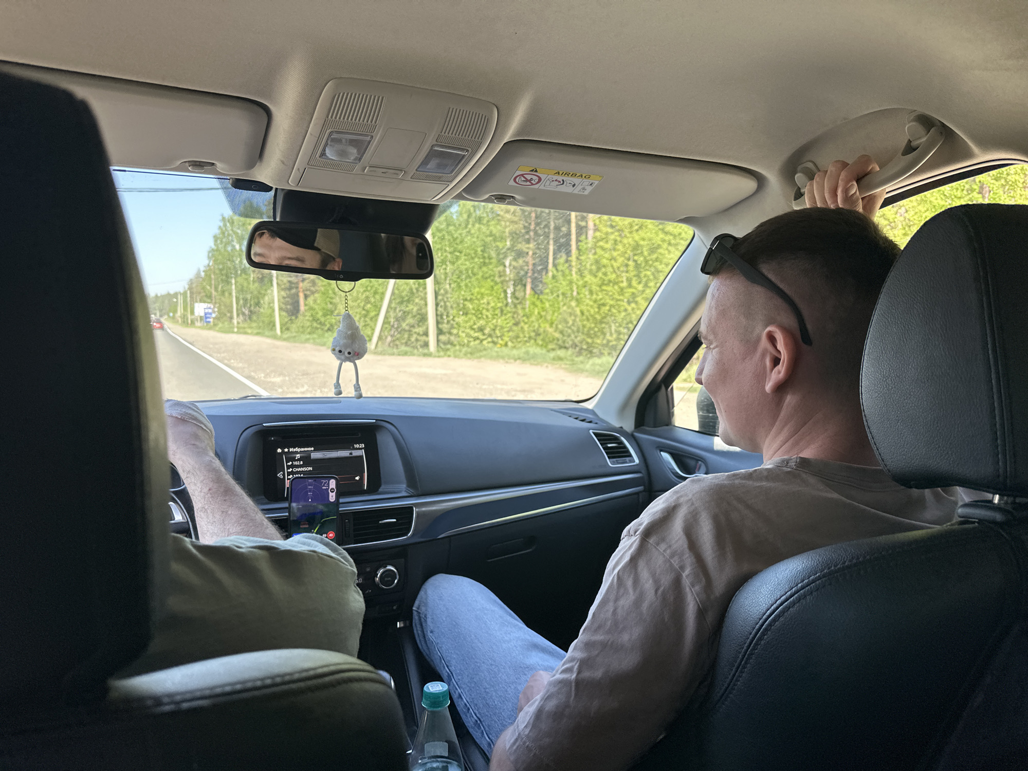 The interior of a car is shown. The vehicle is on a paved road lined with green trees. A person with sunglasses perched on their head is in the passenger seat, holding the car's ceiling handle. The driver's hands are visible, along with a phone mounted on the dashboard displaying navigation. A water bottle is on the floor. The rearview mirror shows the reflection of the road behind them. Based on the environment it is difficult to infer the location of Noviy, Russia.