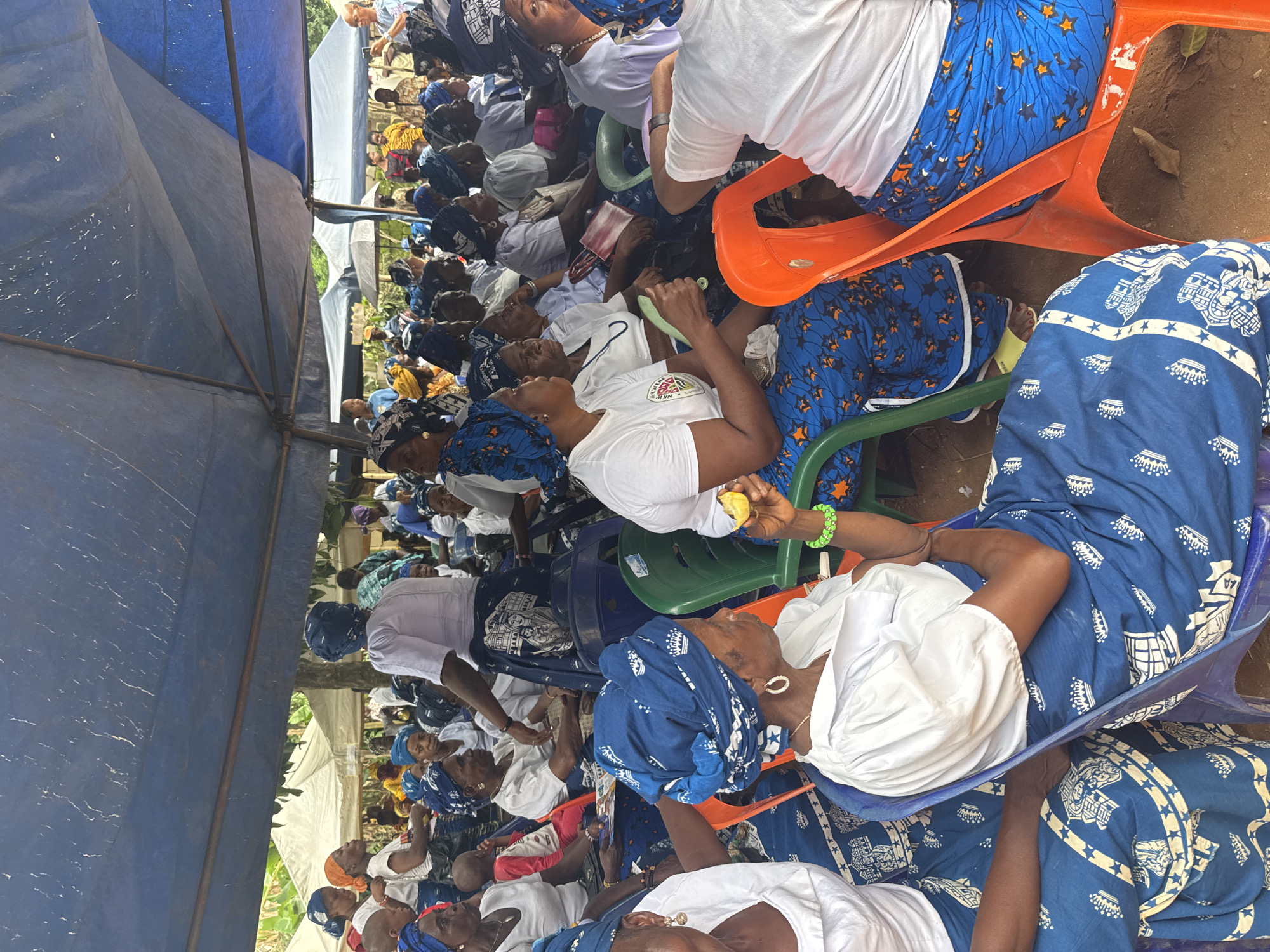 This image captures a vibrant outdoor gathering, likely in Eziudo, Nigeria, under a large blue tarpaulin tent providing shade. A dense crowd of people, predominantly women, are seated in rows on various plastic chairs, including orange, green, and blue ones.
Most attendees are dressed in a coordinated fashion, wearing white short-sleeved tops paired with blue patterned fabrics used as skirts, wraps, and matching headwraps. The patterns on the blue fabric vary, featuring stars, geometric shapes, and stylized motifs.
The individuals appear attentive, generally facing in the same direction, suggesting they are listening to a speaker or participating in a communal event, meeting, or ceremony. In the foreground, one woman holds a piece of fruit, possibly a lemon or lime, and wears a green beaded bracelet. Another woman holds a green, fan-like object. Several women wear jewelry, such as necklaces and hoop earrings.
The ground beneath the chairs is exposed earth. In the background, beyond the main tent, glimpses of other white tents and green foliage can be seen, indicating an open-air or semi-rural setting. The lighting suggests it is daytime, with the tent providing relief from direct sunlight. On one woman's white top, a logo is visible, featuring the text "ALMIGHTY GOD" above a graphic that includes the initials "NKW" or similar, within a multi-colored design.