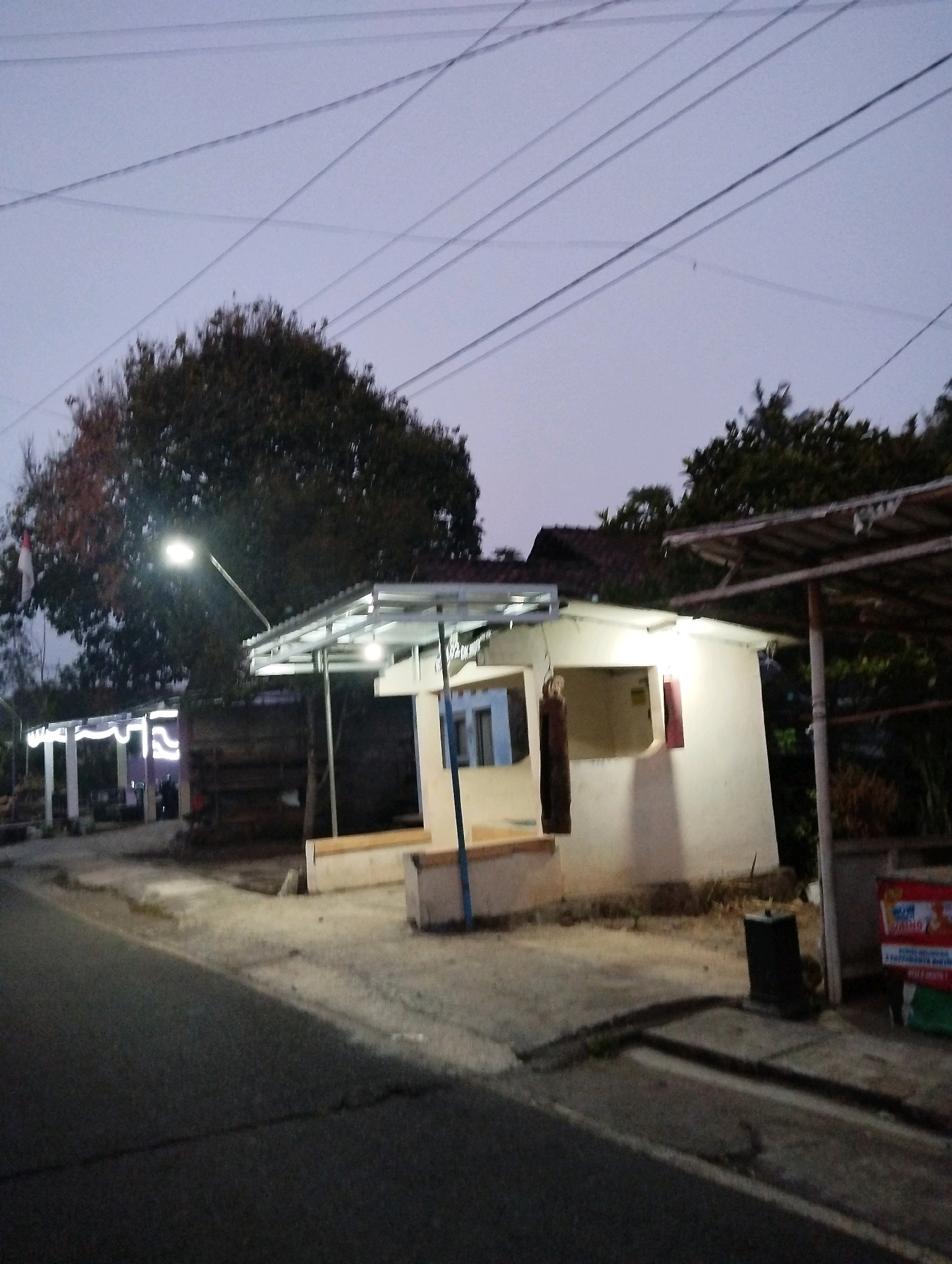 A low-light, outdoor photograph captured at dusk or dawn shows a roadside scene in Salatiga, Indonesia. In the foreground, a dark asphalt road curves to the left. A concrete sidewalk runs alongside the road, bordered by a drainage channel.
To the right of the sidewalk is a small, white, open-fronted building with a corrugated metal roof supported by metal poles. The building has a rectangular opening, suggesting it might be a shop or kiosk. A red-framed window is visible on the side of the building. A dark, rectangular trash receptacle stands near the building.
Further to the left, another structure with a similar corrugated metal roof and white pillars is visible. It is illuminated with decorative string lights. A large tree with dense foliage stands behind this structure, and an Indonesian flag is partially visible on a flagpole to the left of the tree. Electrical wires crisscross the sky above the scene. The overall impression is of a simple, utilitarian roadside setup in a developing urban or semi-urban environment, likely a local marketplace or rest stop.