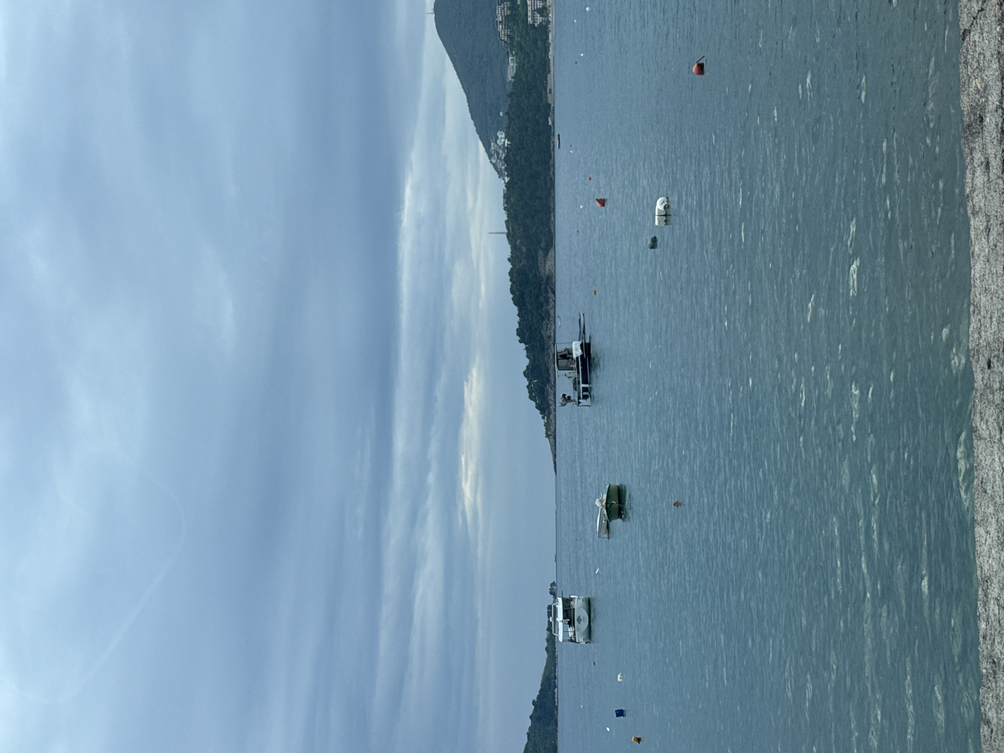 The image shows a body of water with several boats, likely the Adriatic Sea near Sveti Stefan, Montenegro. Along the shore on the right, there is a rocky structure. In the distance, there is a mountainous landmass with vegetation and some buildings visible along the slopes. The sky is cloudy. There are no people immediately visible in the image. Several buoys or markers are also floating in the water. The boats are varying in size and design.