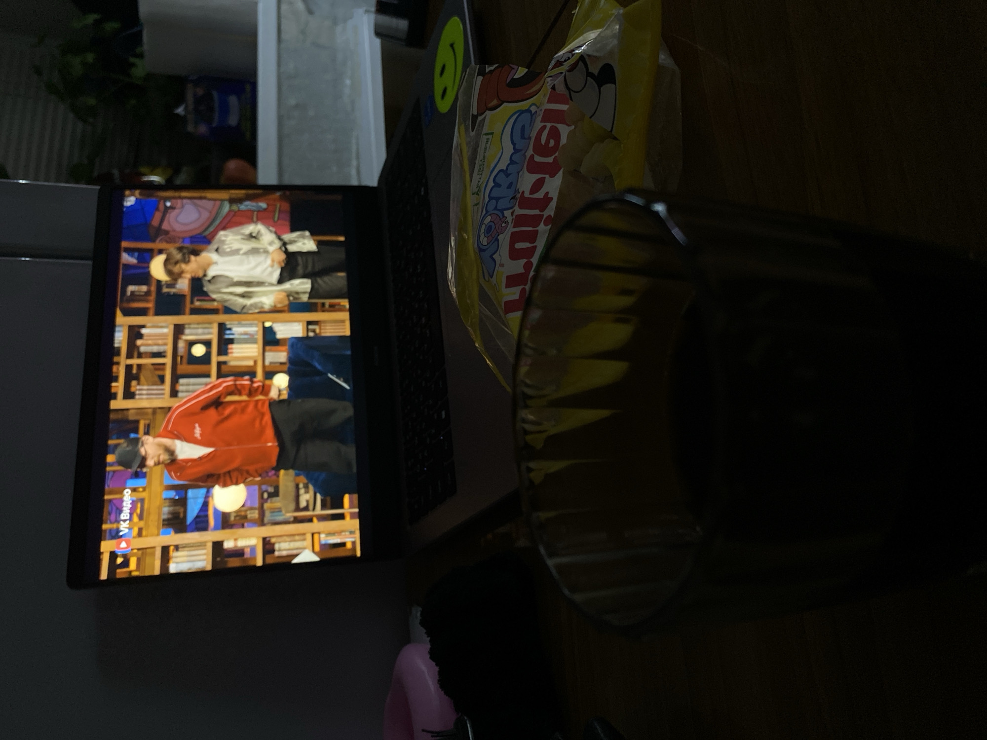 A laptop displays a VK Video of two men standing in front of a bookcase. Adjacent to the laptop is a yellow bag of chips or snacks, and a clear glass. The scene is dimly lit, and there is an object with a pink component towards the lower edge of the photo.