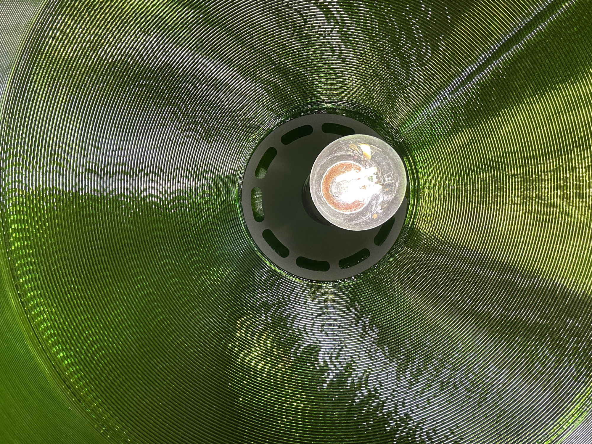A low-angle, indoor shot reveals the interior of a green, ringed lampshade with a lightbulb. The lampshade is composed of concentric rings and textured with minor pitting. The interior is dimly lit, illuminated by the off-center bulb, which is bright with an exposed filament. A dark gray lamp fixture is partially visible, secured around the base of the bulb.
The location is Прикамский, Russia.