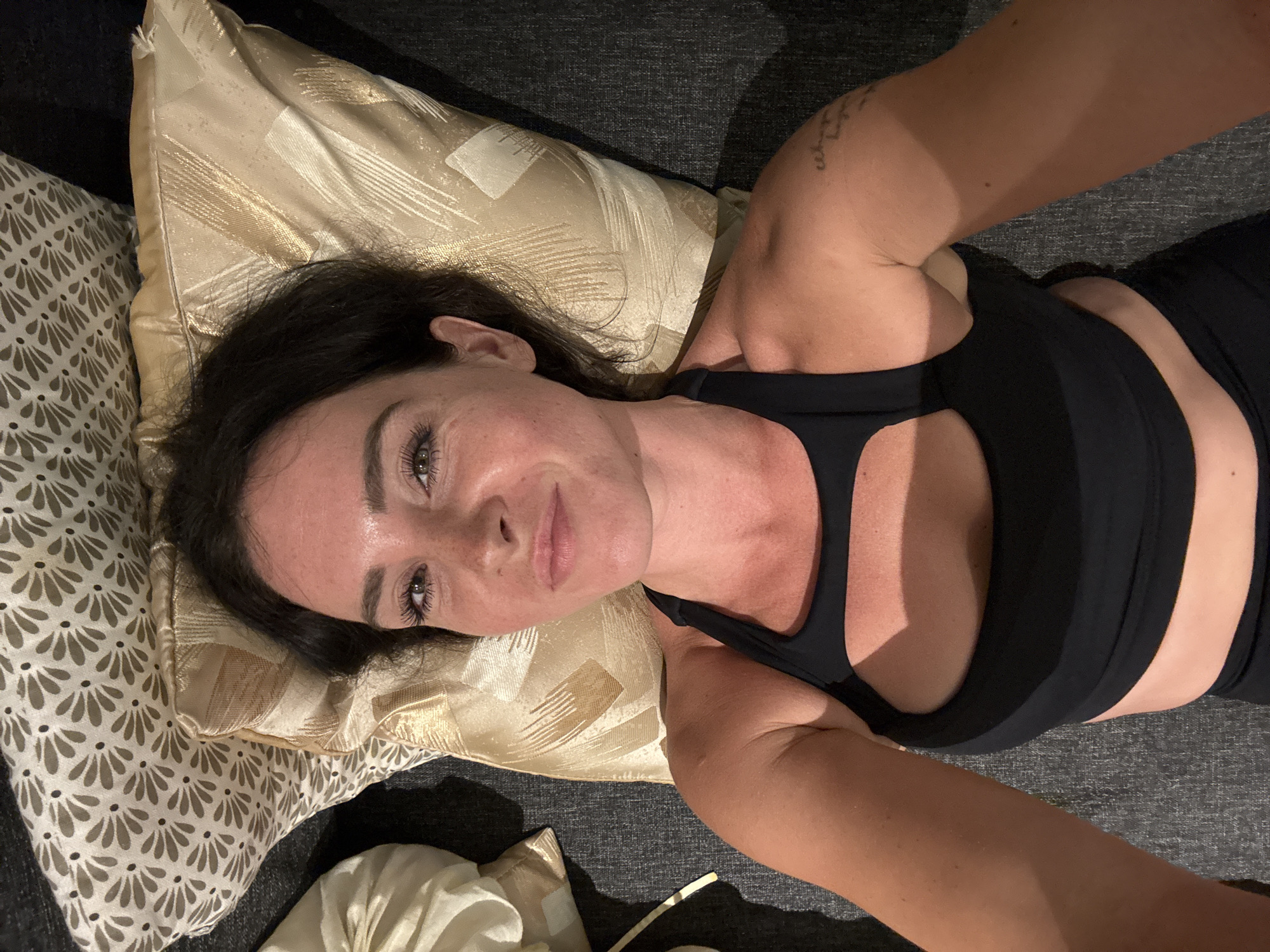 A woman with dark hair lies on her back, propped up by pillows on what appears to be a couch or bed. She is wearing a black sports bra with a cut-out design on the chest. The background consists of a dark gray textured surface, likely upholstery, and several pillows. One pillow behind her head has a floral pattern in light brown and white, while another next to it is a metallic gold color with a textured pattern. A partially visible light-colored pillow is to the left of the frame. The lighting suggests an indoor setting, possibly at night, casting shadows and highlighting the woman's face and shoulders. Her expression is relaxed, and her eyes are open, looking towards the camera. A faint tattoo is visible on her upper right arm. The image is a selfie, taken from a high angle looking down at the woman. This scene is not identifiable as being in North Kuta, Indonesia.