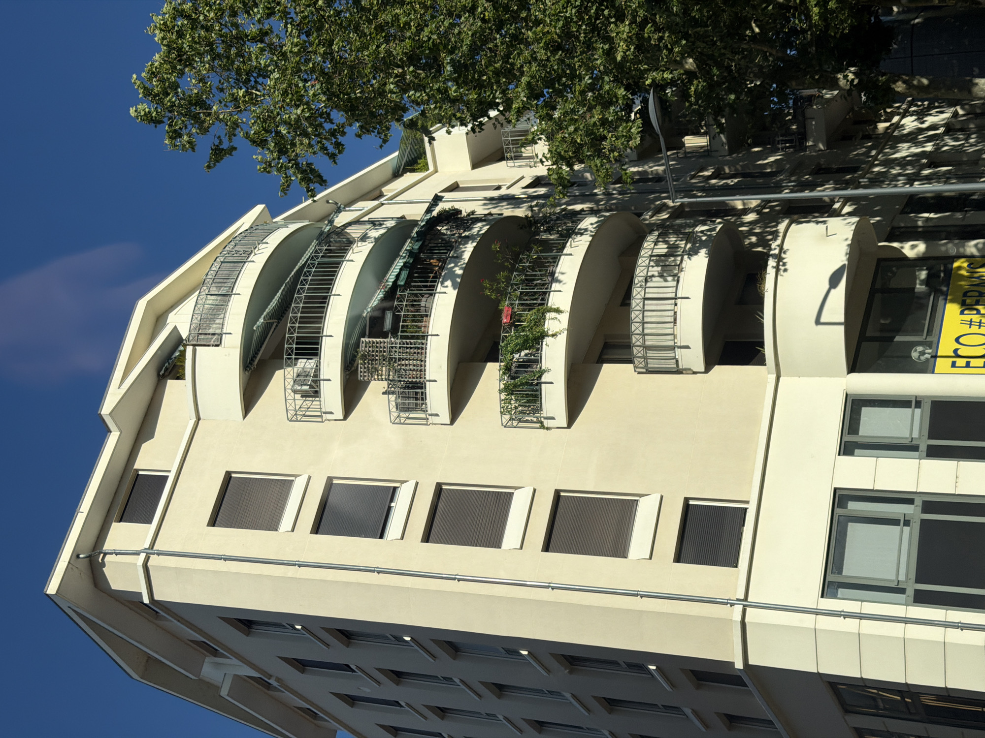 This is a photograph taken from a low angle, looking up at a modern, multi-story apartment building in Lyon, France. The building has a light beige facade and features several curved balconies with metal railings. Some of the balconies have plants and awnings. The building has numerous windows, many of which are covered by dark shutters. The sky is a clear, bright blue. To the right of the building, a large, leafy green tree is visible, casting shadows on the building's facade. In the foreground, a streetlamp stands with its shadow falling across the lower part of the building and the tree. There is a yellow sign with some text on it at the bottom of the building. The architecture suggests a contemporary urban setting.