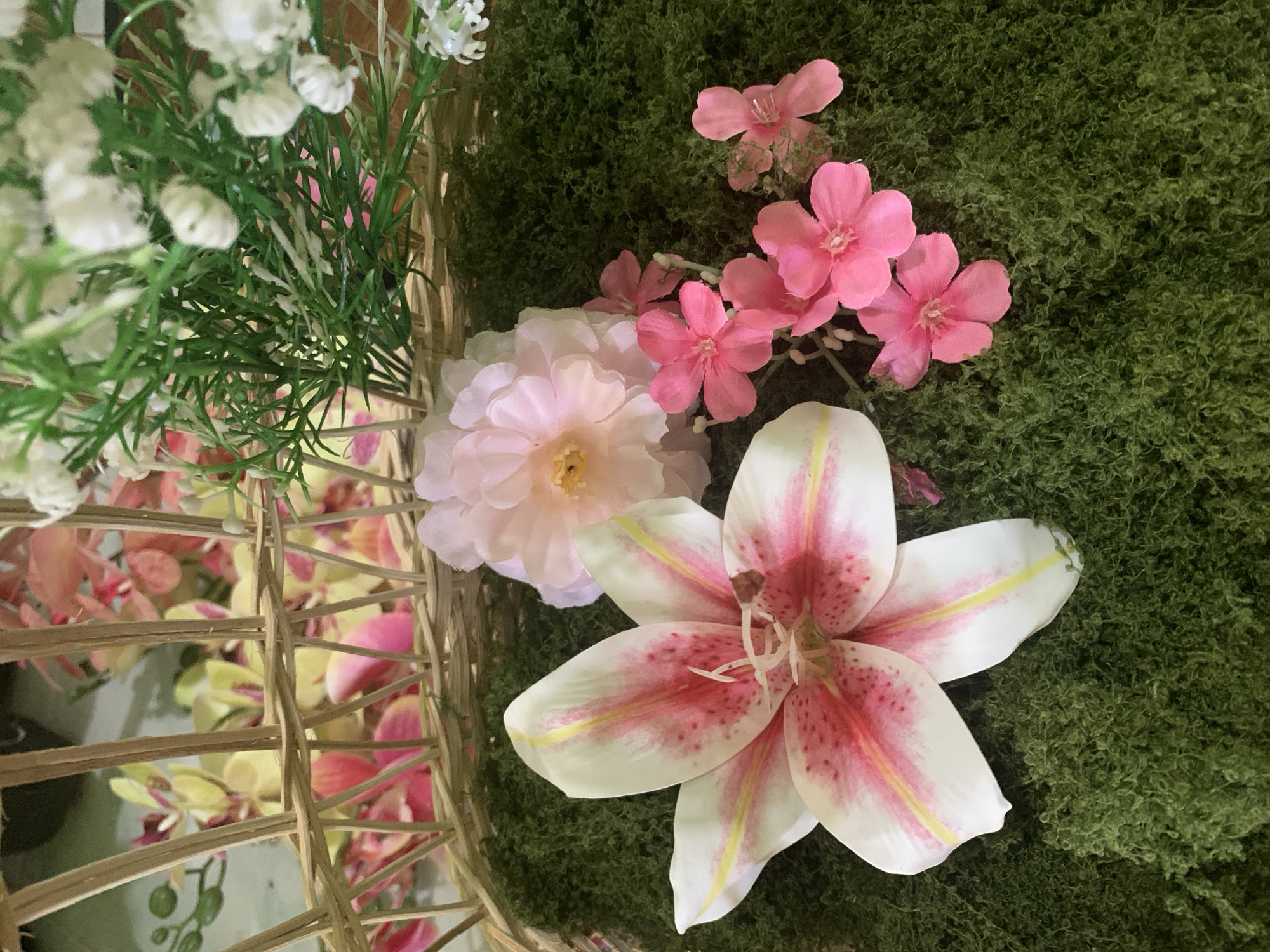 The image displays a close-up of an artificial floral arrangement. The arrangement is contained within a woven basket, which is partially visible in the background. The foreground is covered with a vibrant green, moss-like material, serving as a base for the flowers. Prominently featured is a large, white lily with pink speckles and yellow accents on its petals. To the right of the lily, a cluster of small, bright pink flowers is arranged, accompanied by a larger, pale pink flower behind them. In the upper portion of the image, within the basket, are more artificial flowers, including some with pink and yellow petals, and a sprig of small white flowers with green foliage. The overall scene is a still-life arrangement of artificial flowers, suggesting a decorative or craft-related context. There is no indication of the city of Kuala Pembuang, Indonesia, or any human presence in the image.
