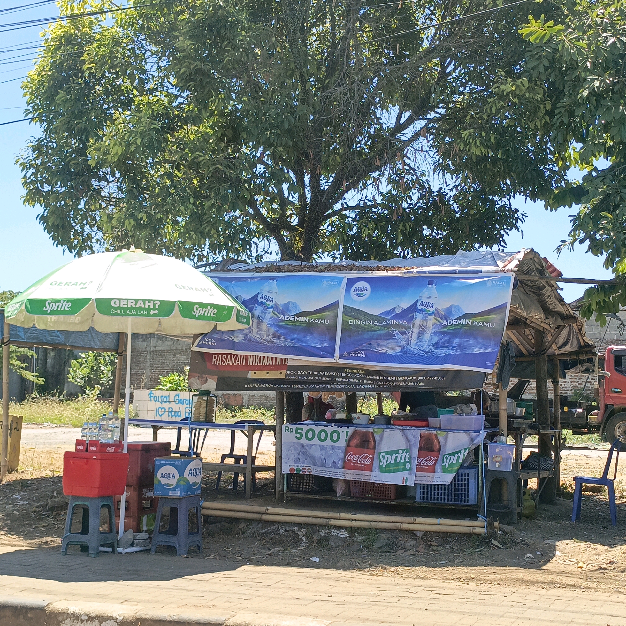 A roadside stall is set up under a large tree in Salatiga, Indonesia. The stall features a white and green umbrella with "Sprite" branding, and a makeshift structure with a corrugated metal roof covered by a tarp. Large banners advertising Aqua water are displayed, with text in Indonesian. In front of the stall, there are various items for sale, including bottled water, Coca-Cola, and Sprite drinks, displayed in red and blue coolers and on tables. A price of "Rp 5000" is visible on a banner. In the background, a red truck is parked. The ground is paved with concrete and dirt. The scene is illuminated by bright sunlight.