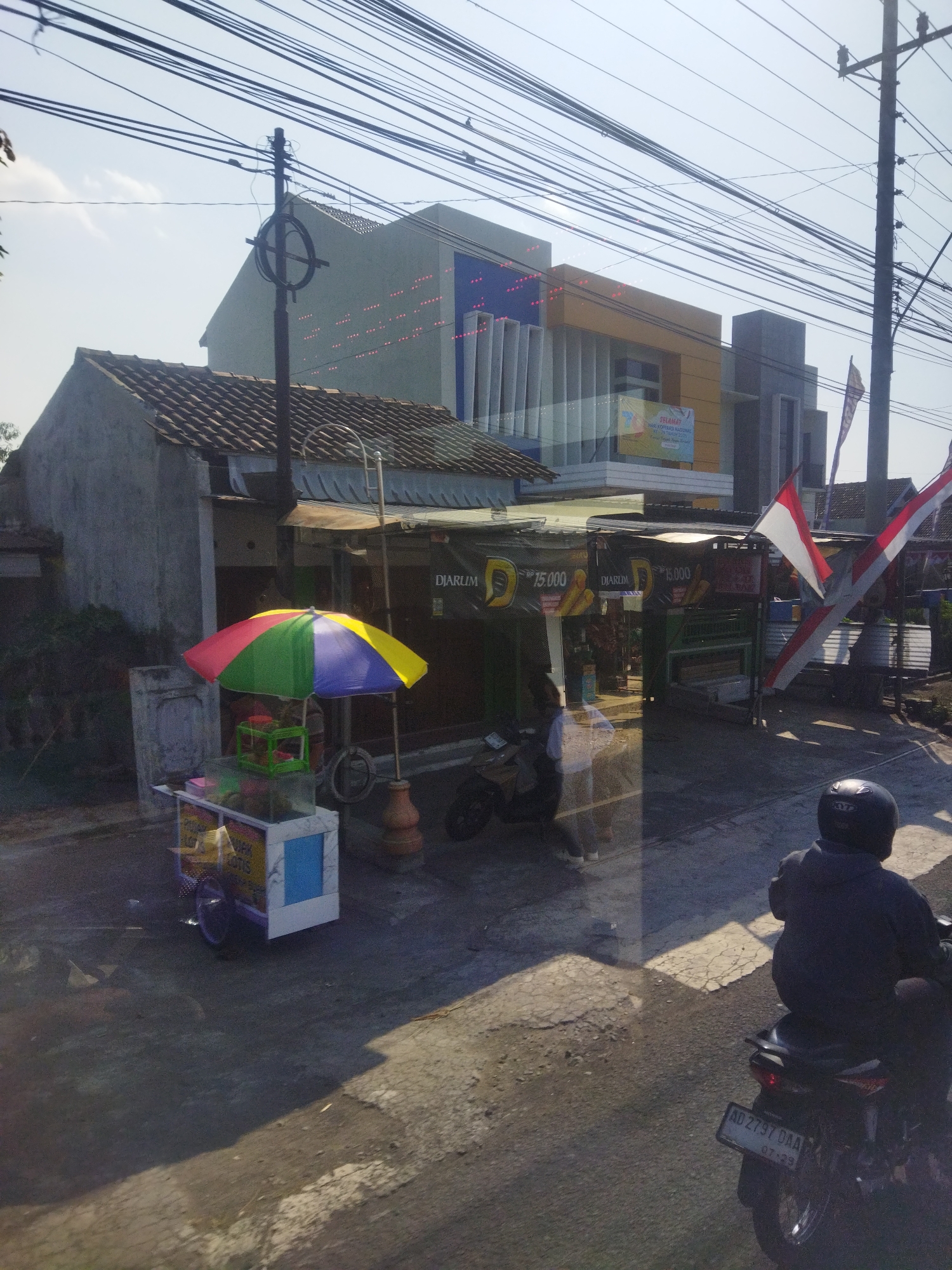 This is a photograph taken in the city of Begajah, Indonesia, showing a street scene. In the foreground, a person is seated on a motorcycle, wearing a dark jacket and helmet. The motorcycle has a license plate that reads "AD 2797 DAA". To the left, a colorful umbrella covers a food cart. The cart has a glass display case containing food items. Behind the cart, a building with a tiled roof and concrete walls is visible. Further in the background, a modern building with yellow and blue accents stands, adorned with a banner that appears to be a congratulatory message. Several Indonesian flags are also present, attached to poles. Overhead, a complex network of electrical wires crisscrosses the sky, supported by utility poles. The ground is paved with asphalt and concrete. The overall lighting suggests it is daytime, with shadows cast by the structures and objects.