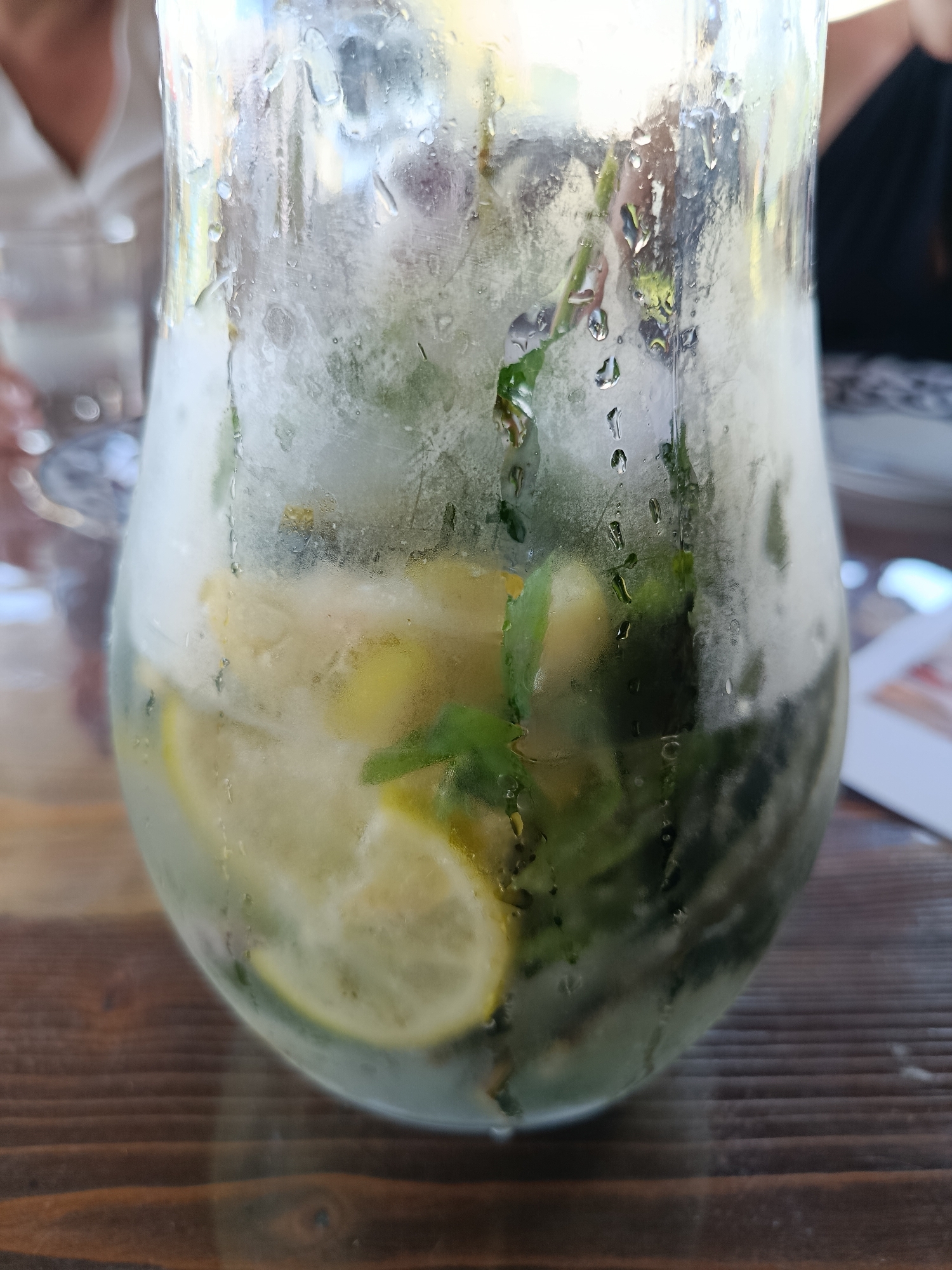 A close-up, eye-level shot captures a condensation-covered pitcher filled with a refreshing beverage, likely a mojito or lemonade, containing sliced lemons and fresh mint. The pitcher rests on a wooden table, with portions of two individuals visible in the background, suggesting an outdoor or casual dining setting. One person, on the left, wears a white shirt, while the other, on the right, is clad in dark attire. The image does not contain information that allows for the determination of its location in the city of Misaylovo, Russia.