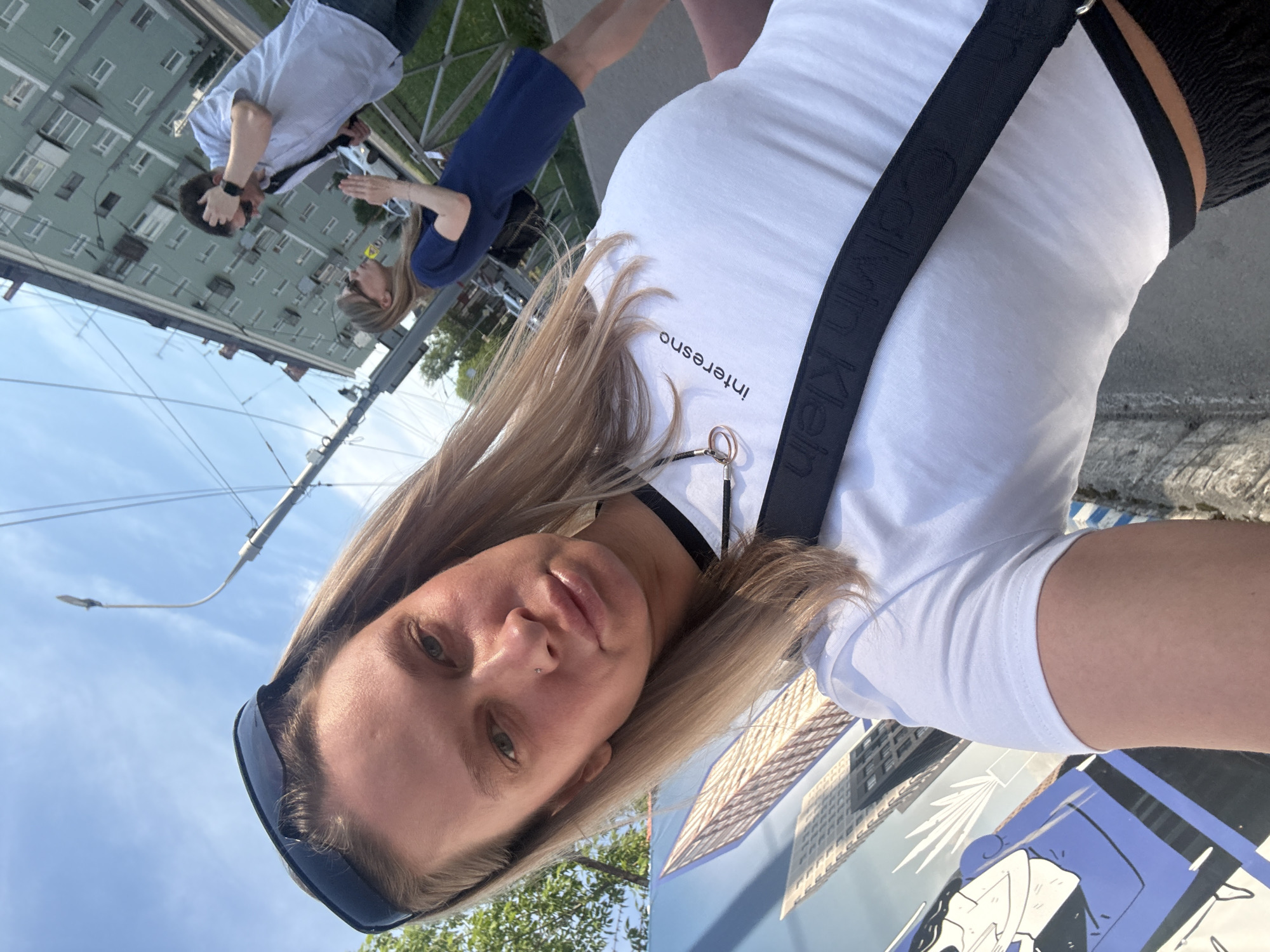 A photograph taken in Perm, Russia, presents a woman in the foreground. She is wearing a white t-shirt with the word "Interesno" printed on it. A black Calvin Klein strap is across her shoulder. In the background, a couple is interacting near a building that matches architectural styles common in Russian cities. The photograph includes a street lamp and hints of trees and blue sky, suggesting an urban outdoor setting. There is an art panel on the ground.