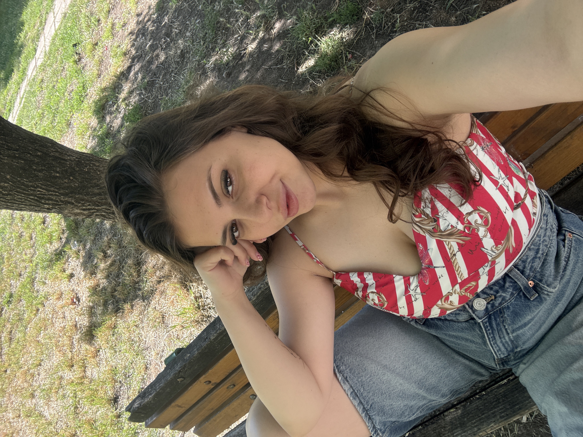 A woman with long, brown hair is sitting on a wooden park bench, looking towards the camera. She is wearing a red and white striped halter top with a nautical print, and denim shorts. Her left arm is resting on her lap, and her right hand is touching her face. The background consists of a tree trunk on the left, and dry grass and dirt with dappled sunlight on the ground. The scene appears to be outdoors, likely in a park, during the daytime. The woman has a relaxed and playful expression. There is no visible text in the image.