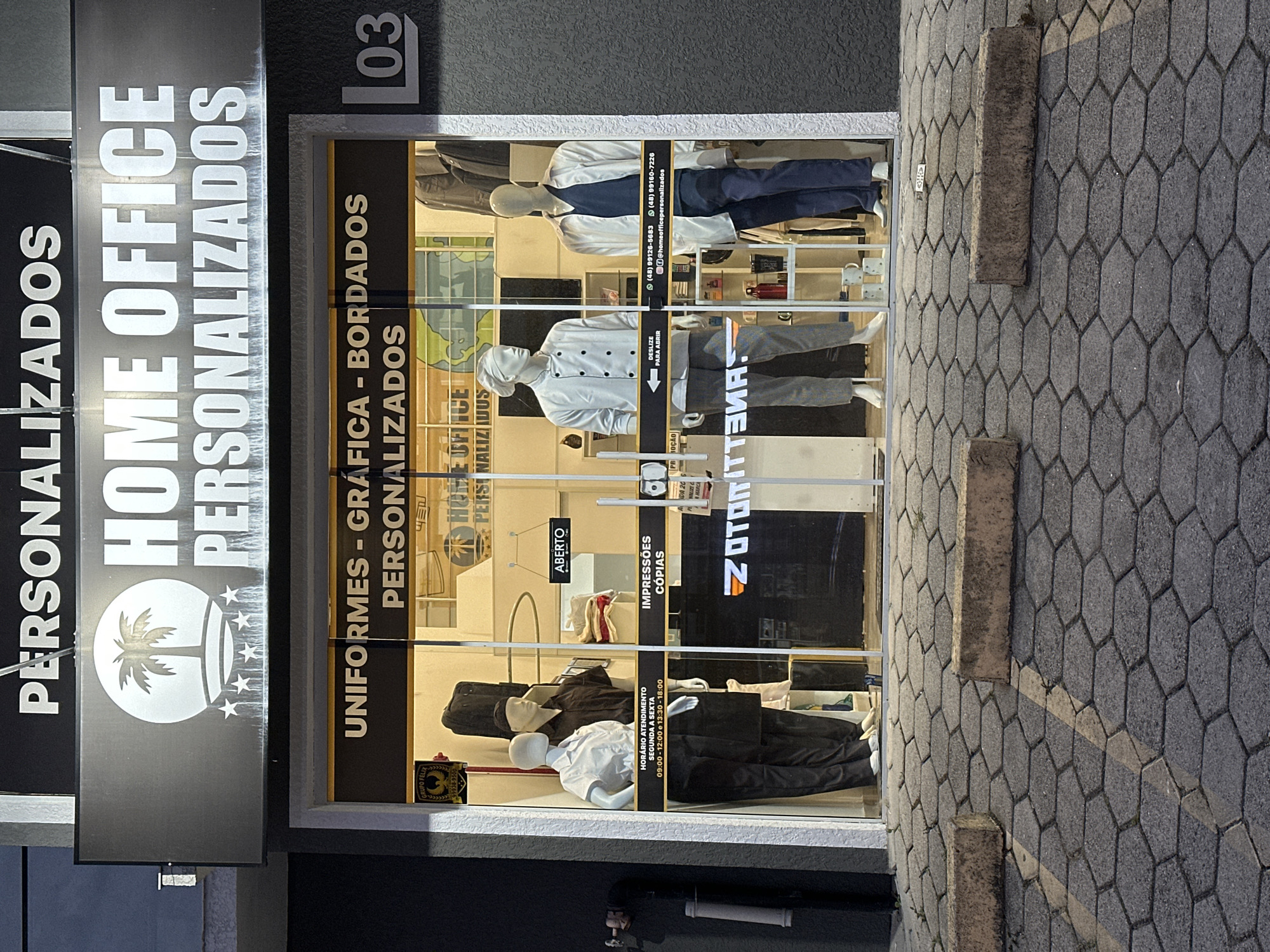 The image depicts the storefront of "Home Office Personalizados" in Florianópolis, Brazil. The sign above the store prominently displays the company's name and logo, featuring a palm tree and stars. The store's window display showcases mannequins dressed in various uniforms, including chef attire and office wear. Information about the store, such as contact numbers and opening hours, is visible on the window. A second sign reads: "UNIFORMES - GRÁFICA - BORDADOS PERSONALIZADOS," also displayed are the words: "IMPRESSÕES CÓPIAS." The store is open, as indicated by the "ABERTO" sign. The address number "03" is indicated on the upper right corner. The sidewalk in front of the store is paved with hexagonally patterned blocks.