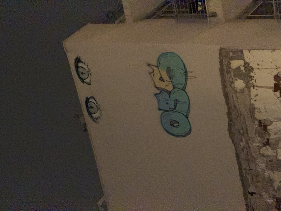The image depicts graffiti on a building exterior. On a gray, concrete wall are two realistically drawn eyes stacked vertically. Further right, a blue and yellow stylized design appears, stacked into three oval-like shapes. To the right, the wall is heavily textured and damaged with crumbling concrete and exposed brick. A cage with metal bars is visible in the upper background. The lighting is dim, obscuring details of the surrounding environment.