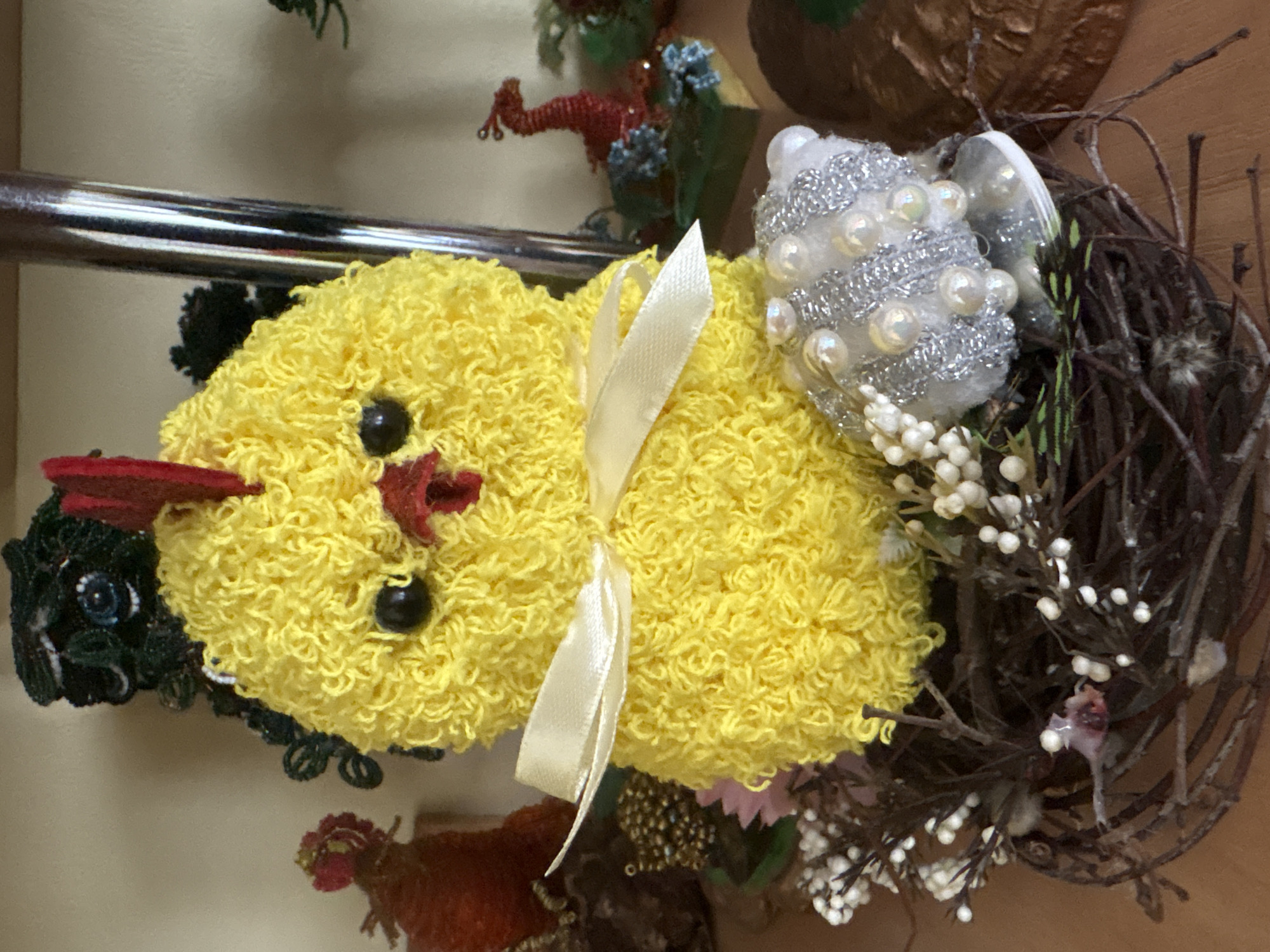 The image shows a close-up of a decorative yellow chick, likely a craft item, situated in a bird's nest made of twigs and adorned with small white berries. The chick has black button eyes, a red comb and wattle, and is wearing a pale yellow ribbon tied in a bow around its neck. To the right of the chick, also partially within the nest, is a small, fuzzy object encrusted with silver thread and white pearls. The background is blurred, but appears to be an indoor setting with other decorative items visible, including what looks like a beaded figure of a rooster to the left and a red beaded dragon to the right. The lighting is warm and appears to be coming from the front. The scene does not depict any specific urban or rural setting within Ветрынскі сельскі Савет, Belarus, but rather a still-life arrangement of decorative objects.