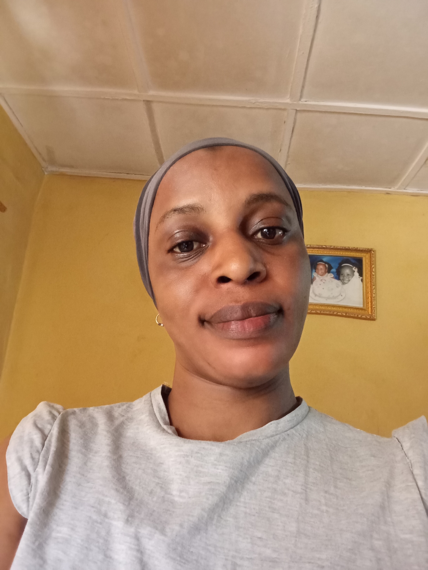 A head-and-shoulders selfie of a woman with medium brown skin, taken indoors. She wears a light grey t-shirt and a dark grey head wrap. She has dark brown eyes, full lips, and a small gold hoop earring in her left ear. The background is a yellow wall with a framed picture hanging on it and a white tiled ceiling. The photo was taken in Narayi, Nigeria.