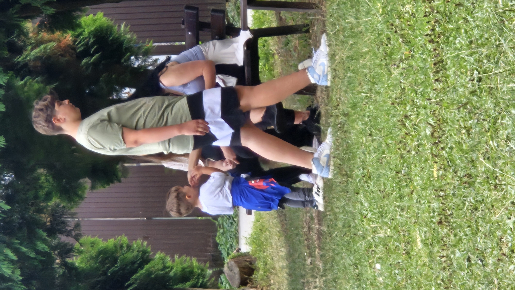 The image depicts an outdoor scene with several individuals, suggesting a public park or garden setting in Prešov, Slovakia, indicated by the trees and grassy area. In the foreground, a young boy in a white t-shirt and grey pants, with a blue garment draped over his shoulders, stands looking to his left. To his right, a taller individual, likely a teenager, is walking away from the camera, wearing a green t-shirt and black and white shorts. Their posture and attire suggest a casual outdoor activity. Behind these two, a woman with dark hair is seated on a wooden bench, partially obscured by the teenager. Her clothing appears to be a light blue top. The background features a wooden fence and lush green trees, characteristic of a temperate climate. The ground is covered in green grass with scattered dry leaves or debris. The lighting suggests it is daytime, likely a sunny or overcast day, with soft shadows. The overall impression is a snapshot of everyday life in an outdoor recreational space within the city of Prešov.