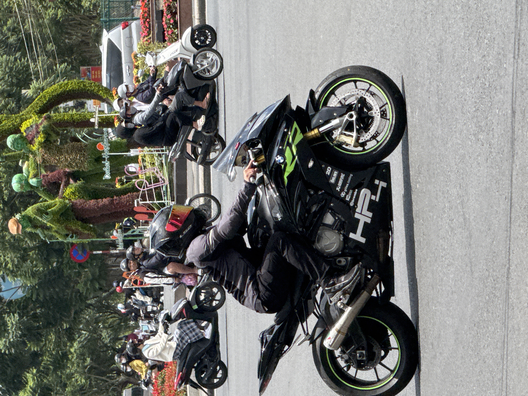 The image depicts a street scene in Da Lat, Vietnam, likely near Xuan Huong Lake, given the presence of lush greenery and floral displays characteristic of the city's tourist areas. In the foreground, a person in black riding gear is on a black and green sport motorcycle. The rider wears a helmet with a red visor and black gloves. The motorcycle features various racing decals, including BMW, HP4, Öhlins, and Brembo. The rider's left foot is on the footpeg, and the right foot is visible near the rear brake pedal.
Behind this motorcycle, several other individuals on scooters and motorcycles are visible. One scooter is ridden by a person in a white jacket and a checkered skirt. Another scooter carries two passengers. Further back, a white scooter is parked. In the background, people are seen on various motorbikes, suggesting moderate traffic.
The environment includes dense green trees, a large, elaborate floral sculpture or topiary in the shape of human figures, and flower beds with red blooms. A white SUV is parked on the side of the road, and a traffic sign is visible. The road surface is asphalt. The overall impression is a sunny day with a vibrant atmosphere, common in Da Lat's popular public spaces.