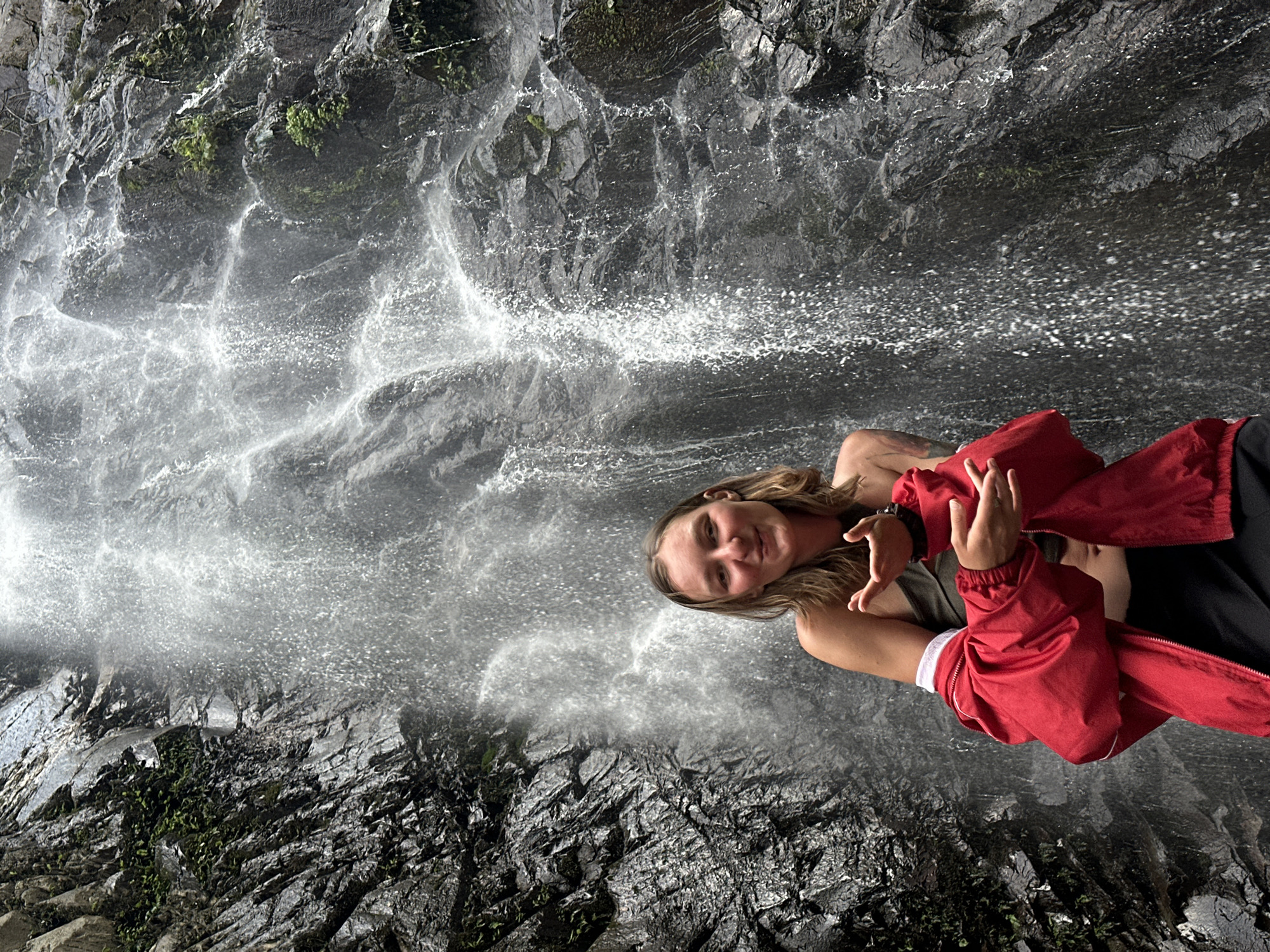 The image captures a young adult female posing in front of a waterfall in Tu Le, Vietnam. The subject, positioned centrally, has light-colored skin, long light brown hair, and is looking directly at the viewer with a smile. She is wearing a dark olive green strapless top, a bright red long-sleeved jacket unzipped and worn off her shoulders, exposing her midriff, and dark-colored bottoms. Her hands are held in front of her chest, with fingers loosely curled and palms slightly open, as if in an expressive gesture.
Behind the woman is a large, cascading waterfall flowing over a dark, rugged rock face. The rock is dark grey to black, appears wet and textured, and is interspersed with patches of green foliage, including ferns and small plants, particularly visible on the left side of the frame and in smaller clusters throughout the rock face. The waterfall itself consists of numerous streams and sprays of white water, creating a dynamic and misty background. The upper portion of the waterfall and cliff extends out of the frame. The overall scene depicts a natural, outdoor environment.