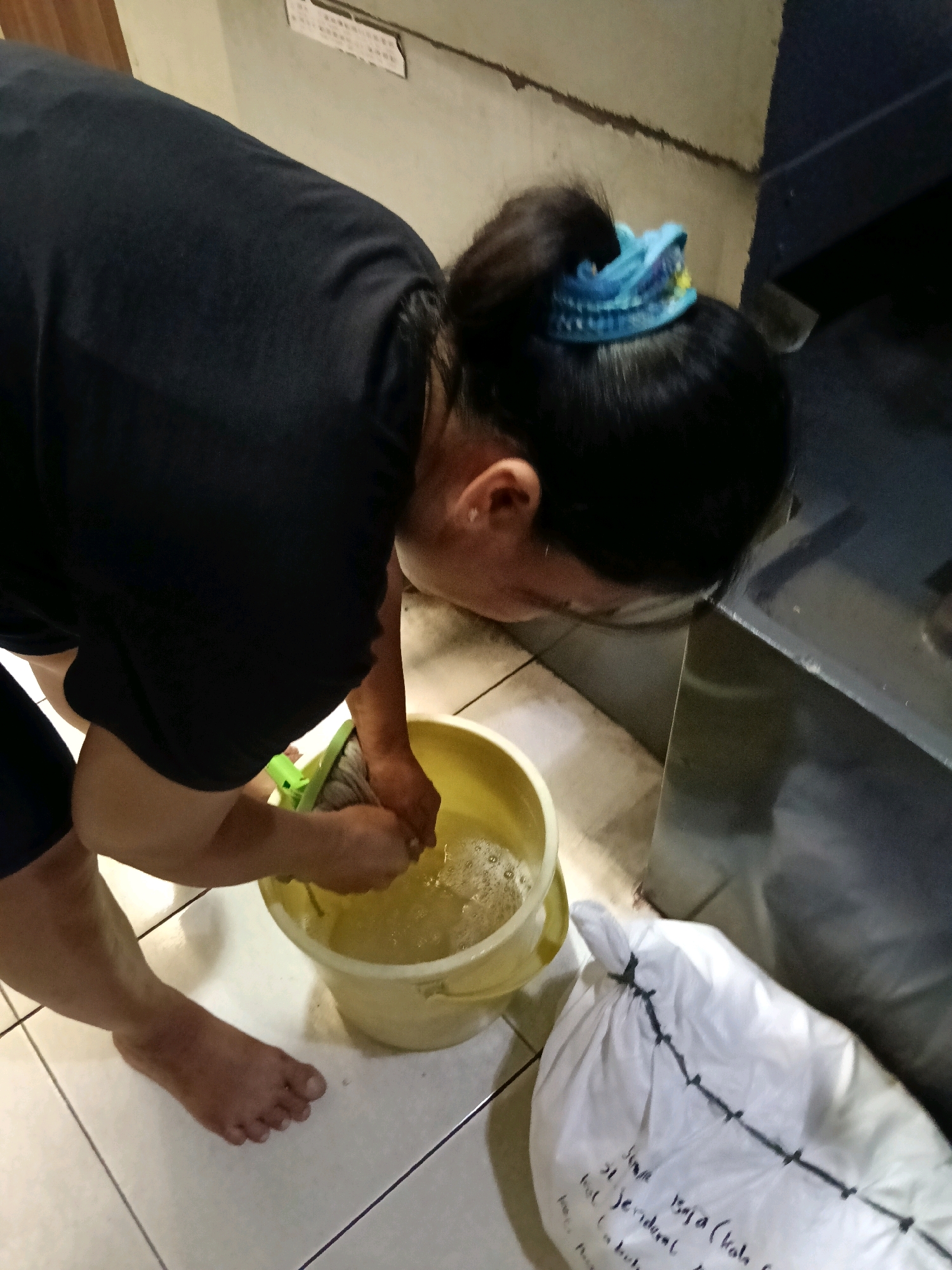 In the city of Belimbing, Indonesia, a person with dark hair tied back with a blue cloth is washing a mop in a yellow bucket filled with soapy water. The individual is wearing a black t-shirt and dark shorts. Their left foot is bare and resting on a white tiled floor. To the right of the bucket, a large white plastic bag is visible, with black markings and writing on it. The background shows a light-colored wall and a dark metallic surface.