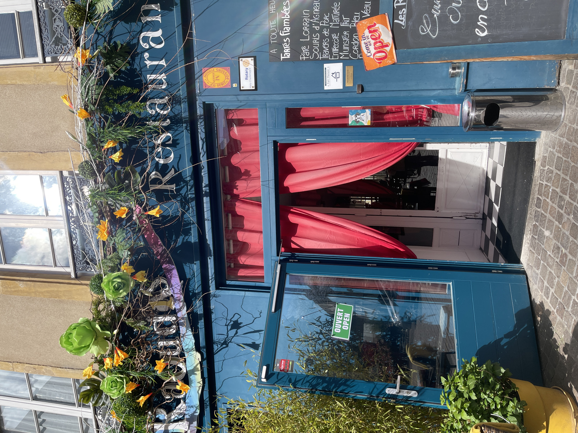 This is a photograph of the exterior of a restaurant in Metz, France. The restaurant's name, partially visible, is "Restaurant." The façade is decorated with flowers and greenery. The entrance features an open blue door, red curtains, and a sign indicating "OUVERT/OPEN". A chalkboard displays menu items in French, including "Tartes flambées," "Pâte Lorrain," and "Cordon Bleu." Various signs and stickers are affixed to the door and walls, including those referencing "Rotary Metz Charlemagne," "Fait Maison," and "Negroni Club." An "Open" sign invites customers inside. A metal ashtray stands next to the entrance. The building's exterior has a stone and stucco finish.