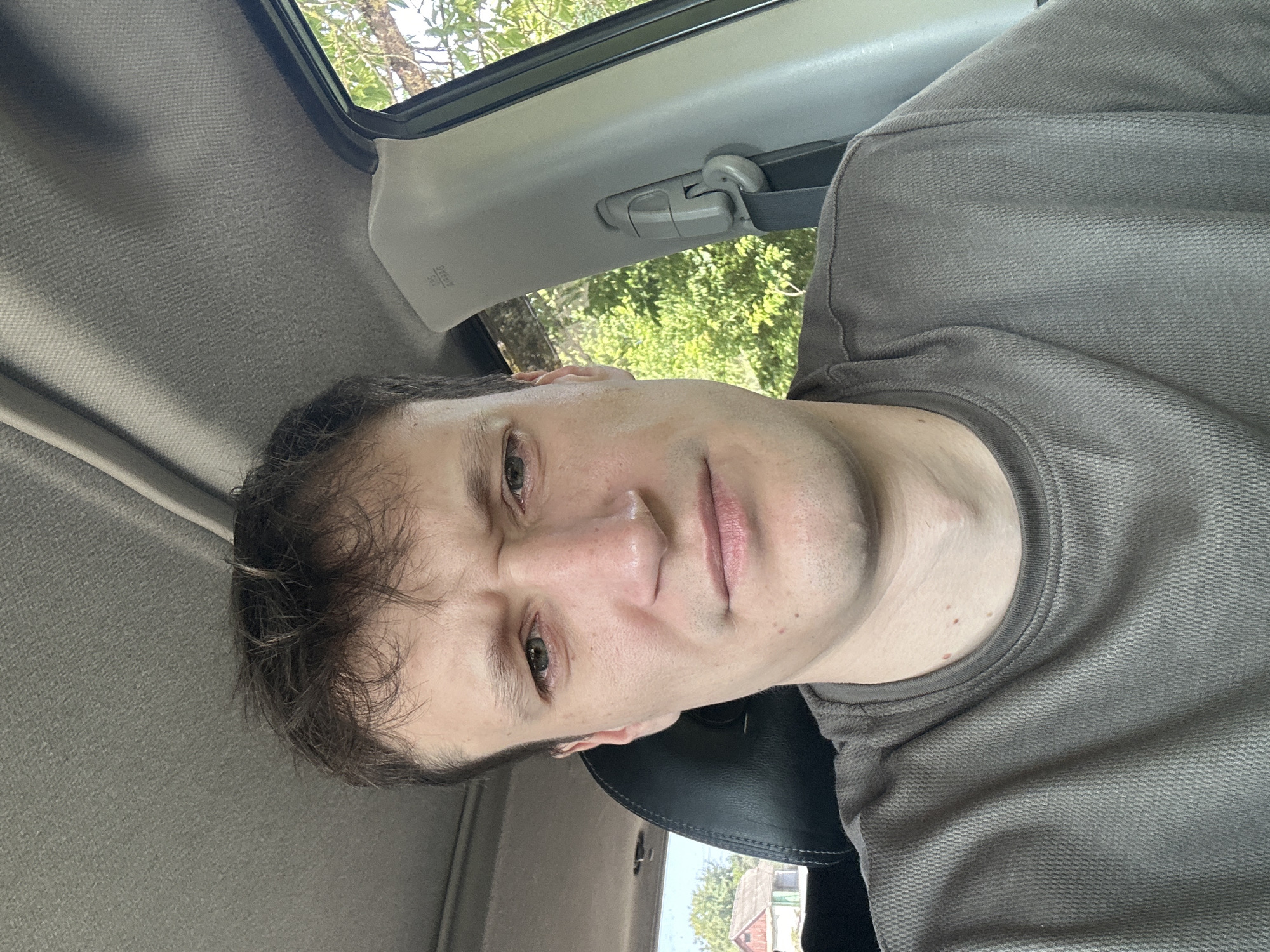 This is a close-up, self-taken photograph of a man sitting inside a car. He has light skin, brown hair, and is wearing a grey t-shirt. His facial expression appears concerned or displeased, with his eyebrows furrowed and mouth slightly pursed. The interior of the car is visible, including the grey fabric headliner and a black seat. Through the open window to the right, lush green trees and foliage are visible, suggesting an outdoor setting. A glimpse of a house with a red roof can be seen to the left. The photograph appears to be taken during daylight. The context of Makiv, Ukraine is not ascertainable from the visual information provided in the image.