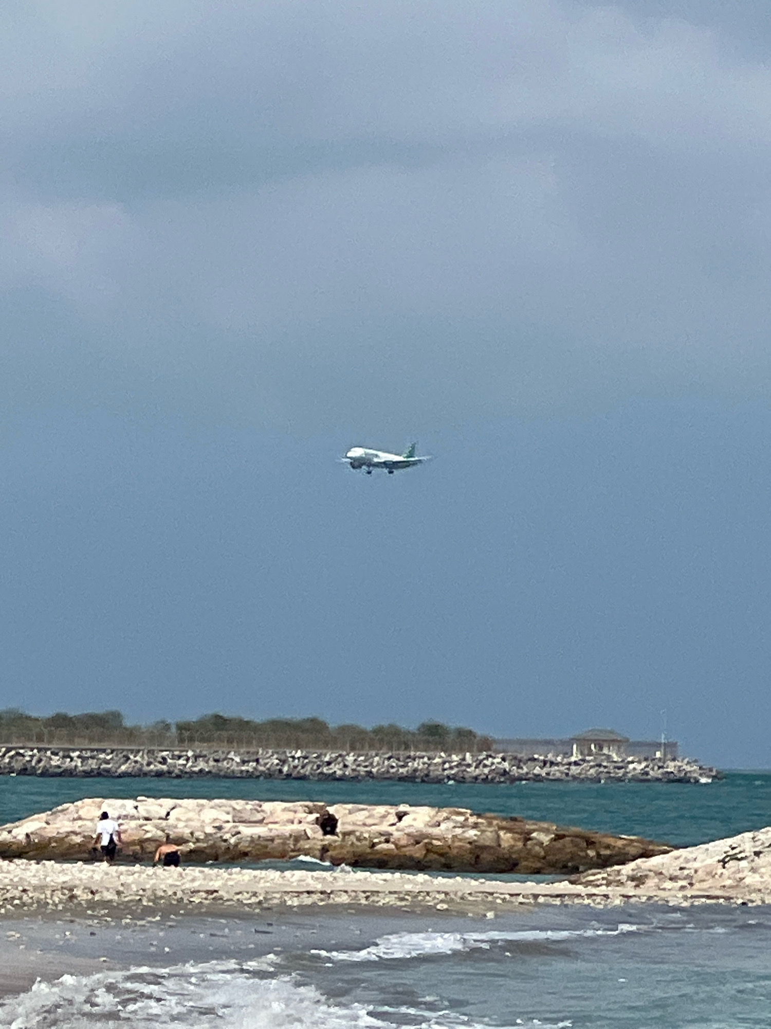 An airplane with green accents is visible in the sky above a body of water. In the foreground, waves crash on a pebble beach. A man in a white shirt and black shorts stands on a rocky breakwater, while another person kneels on the same structure. Further in the background, a long, rocky breakwater stretches across the turquoise water, with trees and a building visible on the shore beyond. The sky is overcast with a muted blue hue. The scene appears to be a coastal area.