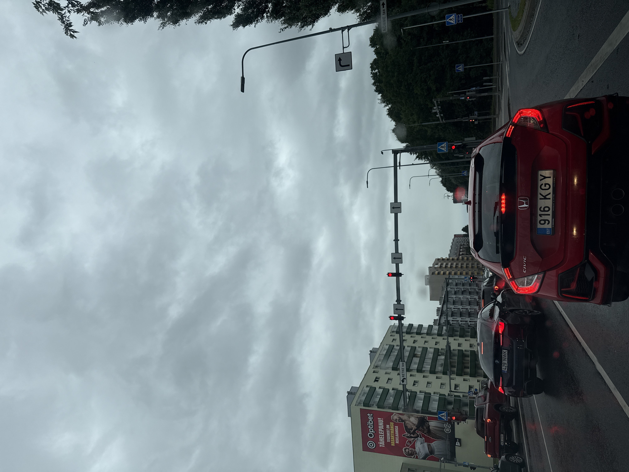 The image depicts a cloudy day in Tallinn, Estonia, likely on a street with moderate traffic. In the foreground, a red Honda Civic is the most prominent subject, facing away from the camera, with its brake lights illuminated. To its left, a dark colored BMW sedan, license plate 429 NWK, is also in traffic. Further left, a red pickup truck with license plate 435 XYM is visible. The road appears wet, indicating recent or ongoing rain.
To the left of the road, a multi-story building is prominent, featuring a large advertisement for "Optibet". Across the street, other buildings and traffic lights are visible. Traffic lights are present at multiple intersections, with some indicating red. A directional sign points to "RETKE tee". On the right side of the road, trees form a dense line, and a sign for "NÕMME tee" is visible. The sky is overcast with heavy, grey clouds. The overall atmosphere suggests a typical urban environment during inclement weather.