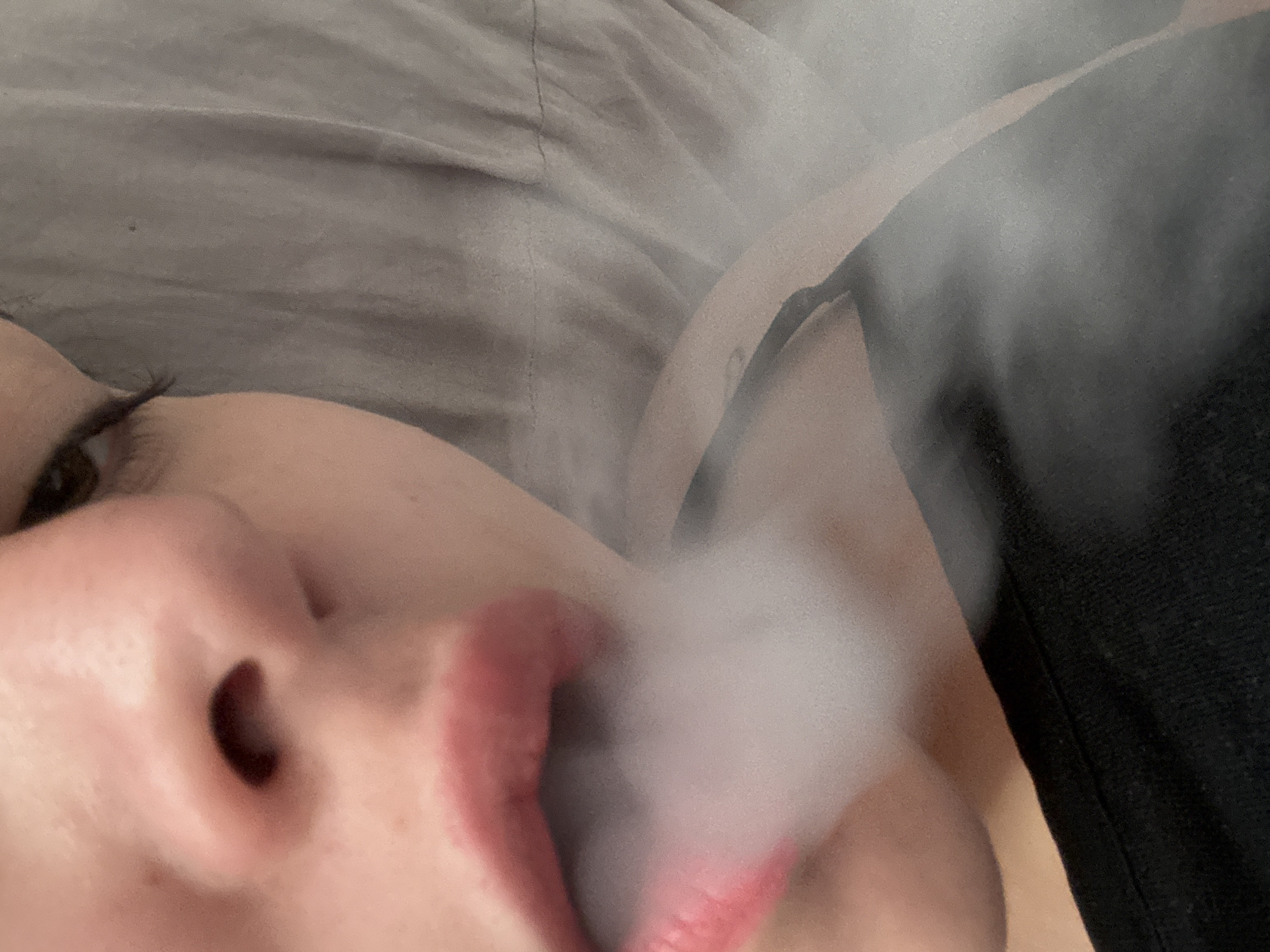 The image shows a close-up of a person's face exhaling smoke. The person has fair skin, dark eyelashes, and red lipstick. The smoke obscures part of their face. They are wearing a black top. The background is a grey, textured surface, possibly a bedsheet. The image does not appear to be taken in Troyan, Bulgaria. No location is identifiable in the image.