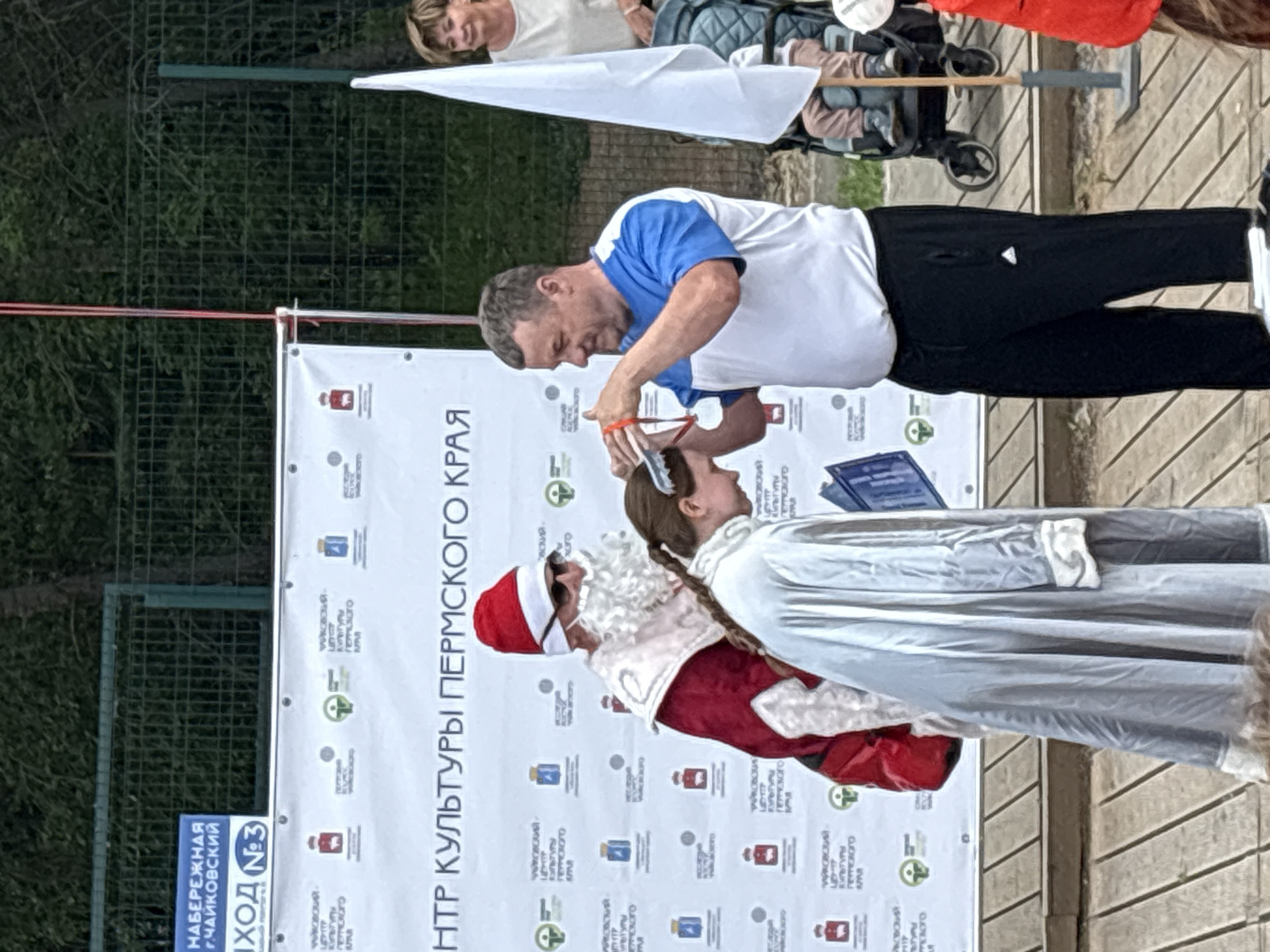 This media file shows an outdoor event in Chaikovsky, Russia. The main subjects are three people: a man in a blue and white shirt, a young girl dressed in a silver costume, and a person in a Santa Claus costume. The man appears to be cutting the girl's hair or performing a similar action with scissors near her head. The Santa Claus figure is standing next to them, looking towards the girl.
Behind the figures is a large white banner with Cyrillic text and various logos. Text visible on the banner includes "ЦЕНТР КУЛЬТУРЫ ПЕРМСКОГО КРАЯ" (Center of Culture of Perm Krai). There is also a blue sign with text that includes "НАБЕРЕЖНАЯ Г.ЧАЙКОВСКИЙ" (Chaikovsky Embankment) and "ВХОД №3" (Entrance No. 3).
In the background, there is a green fence and some trees. There are also glimpses of other people and a stroller, suggesting this is a public event or gathering. The lighting suggests it is daytime, possibly overcast. The overall scene appears to be festive and community-oriented, possibly related to a cultural event or a holiday celebration.