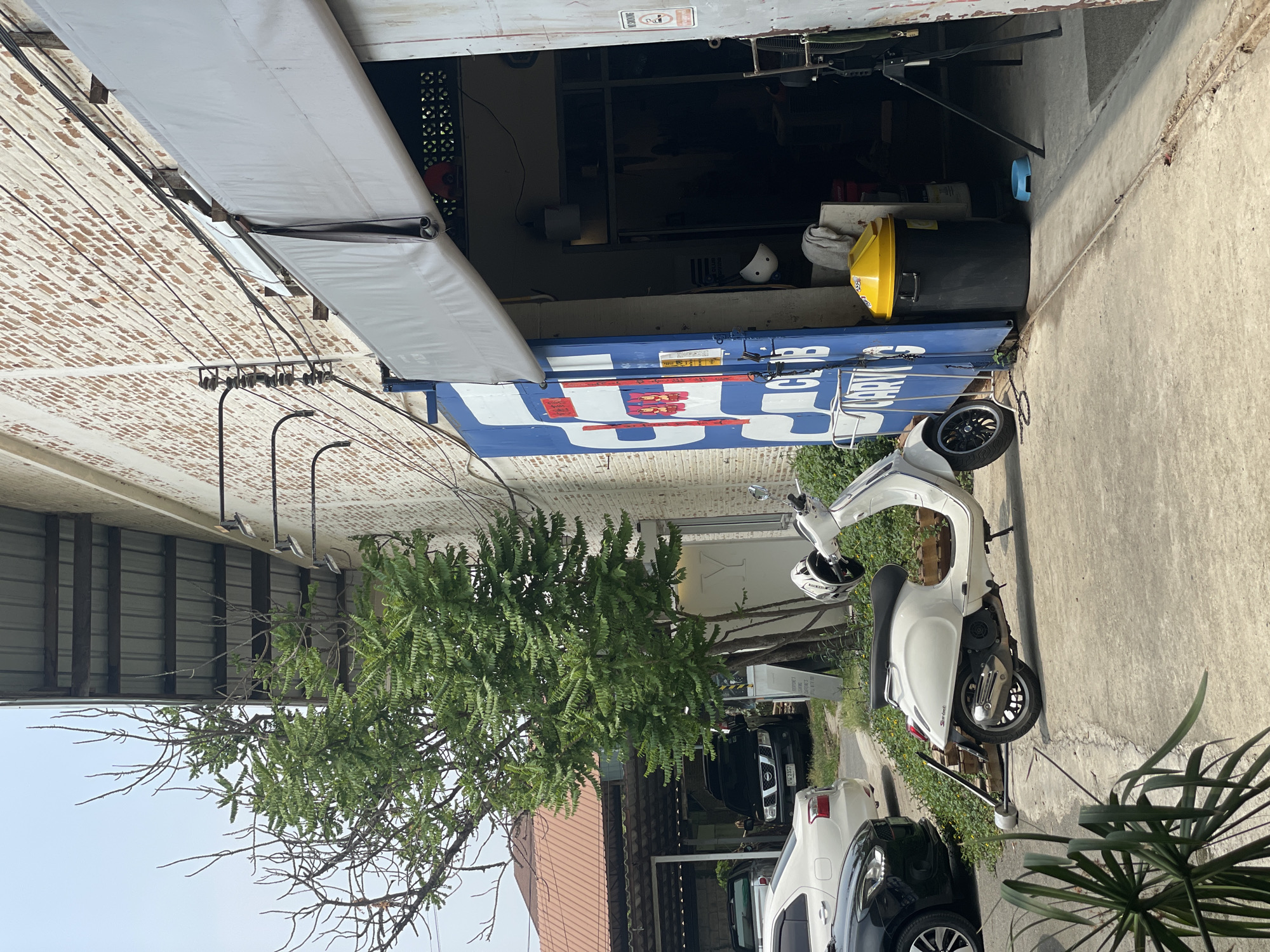 The image shows a street scene in Ban Rong Ruea Kham, Thailand. A white scooter is parked on the sidewalk, next to a black car and a white car. A tree partially obscures the view of a building behind the vehicles. A large blue and white container, possibly repurposed, is positioned near the street, its side marked with writing and a logo. An awning extends from the building. There is a trash can and a helmet visible inside of the repurposed container. Power lines are strung across the frame.
