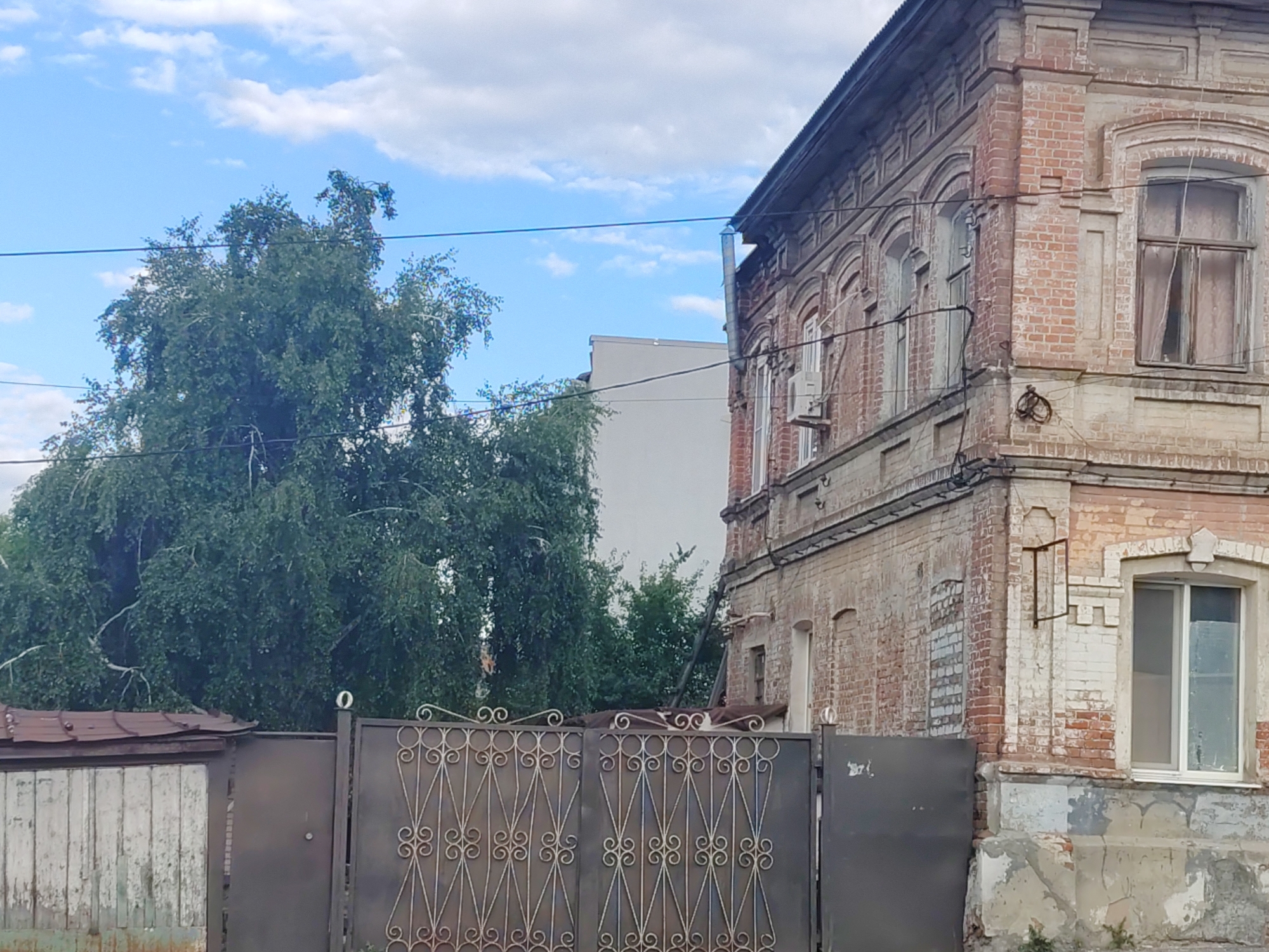 The image depicts an exterior scene in Saratov, Russia, dominated by an old, ornate brick building on the right and a large, leafy green tree on the left. The building features arched windows and decorative brickwork, showing signs of age and weathering. A metal gate with intricate scrollwork is visible in the foreground, partially obscuring a weathered wooden structure. The sky above is blue with scattered white clouds. The overall impression is one of a somewhat aged urban environment.
