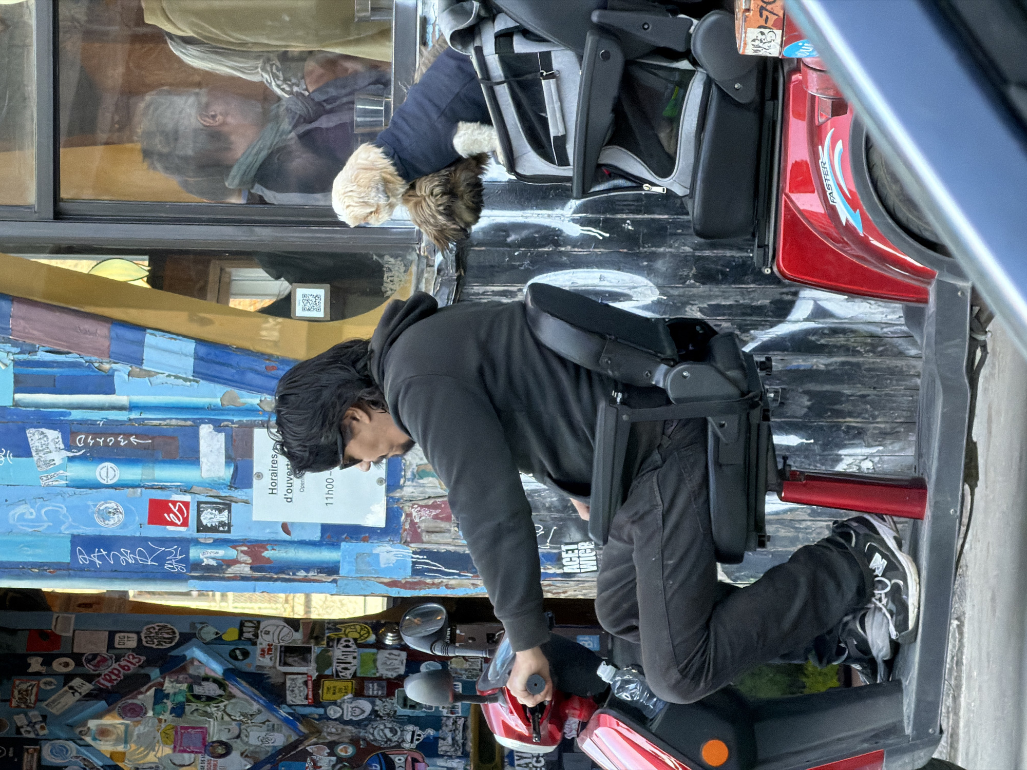 A man in a black hoodie and pants sits on a red scooter in front of a wall covered with stickers and painted blue planks in Montreal, Canada. The wall also features a sign indicating "Horaires d'ouvert/Open 11h00-". A dog is visible in a carrier attached to the back of the scooter. Reflected in a nearby window is another person wearing a grey scarf and jacket.