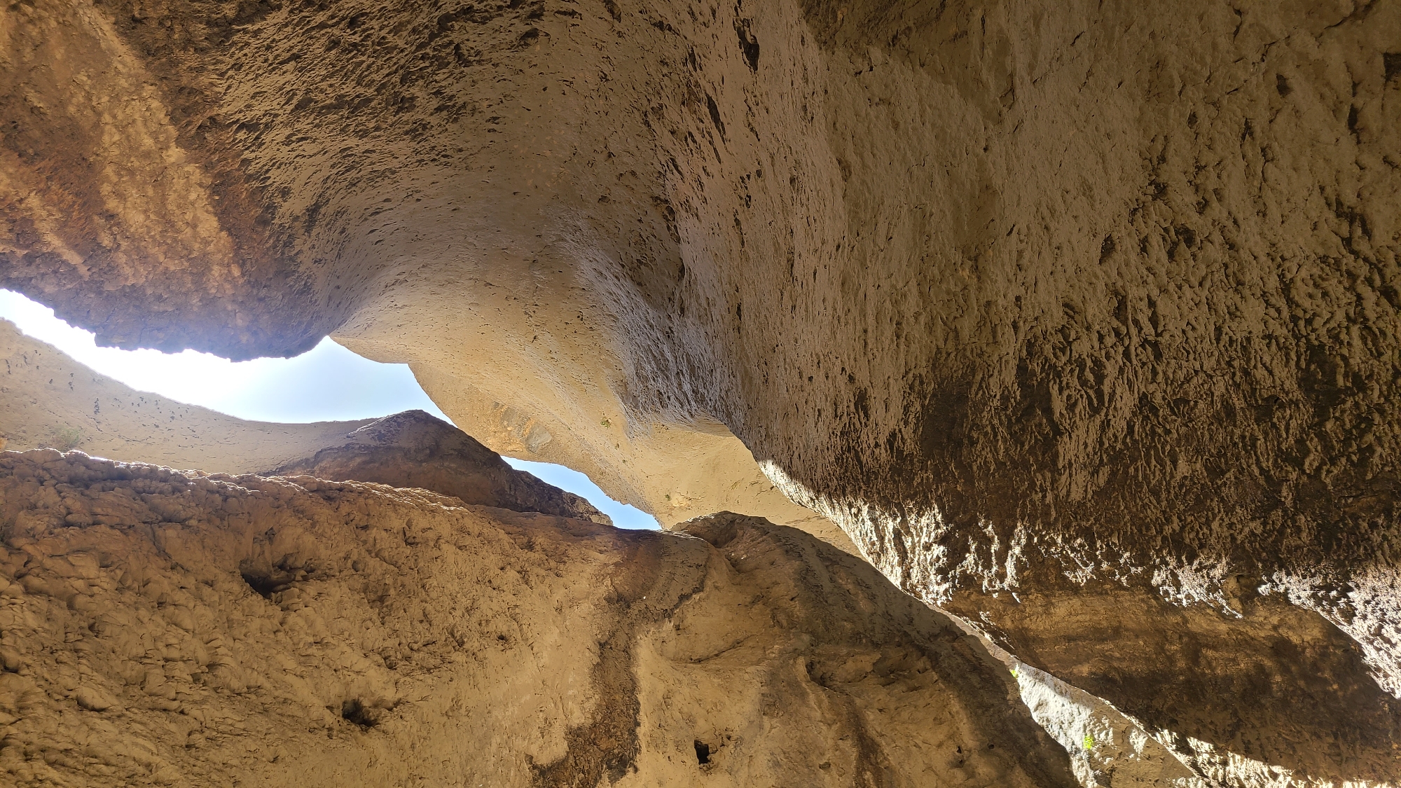 The image depicts a view looking upwards within a natural rock formation in the city of Ziarat, Pakistan. The scene shows a tight space between large sandstone-colored rock formations, possibly inside a cave or narrow canyon. Sunlight is visible through gaps in the rock ceiling, revealing a blue sky. The surfaces of the rocks are textured, with some showing evidence of weathering and erosion. There are no people or identifiable objects present. The overall impression is of a natural, geological feature.