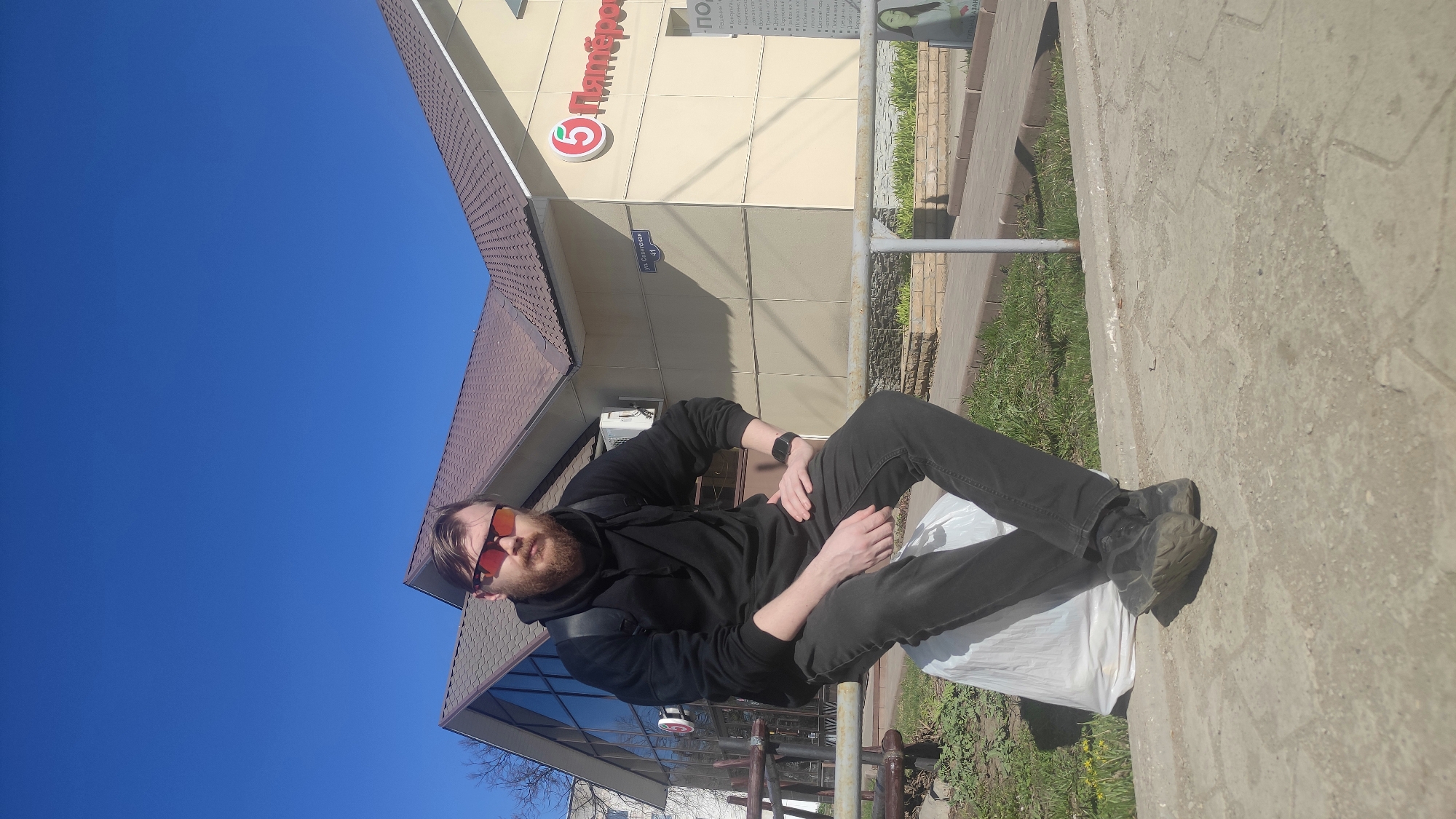 A full-body shot shows a man with a beard and red sunglasses sitting outdoors with his legs crossed. He's wearing a black hooded sweatshirt, dark pants, and dark shoes. He appears to be sitting on a white bag. The location is Chaikovsky, Russia. Behind the man is a building with a sign in Russian that reads "5 Пятёрочка" (Pyaterochka), a grocery store chain. A street sign reads "ул. Советская 41" (Ulitsa Sovetskaya 41), indicating the building's address. The sky is blue, and the photo seems to have been taken on a sunny day. The man is wearing a watch on his left wrist.